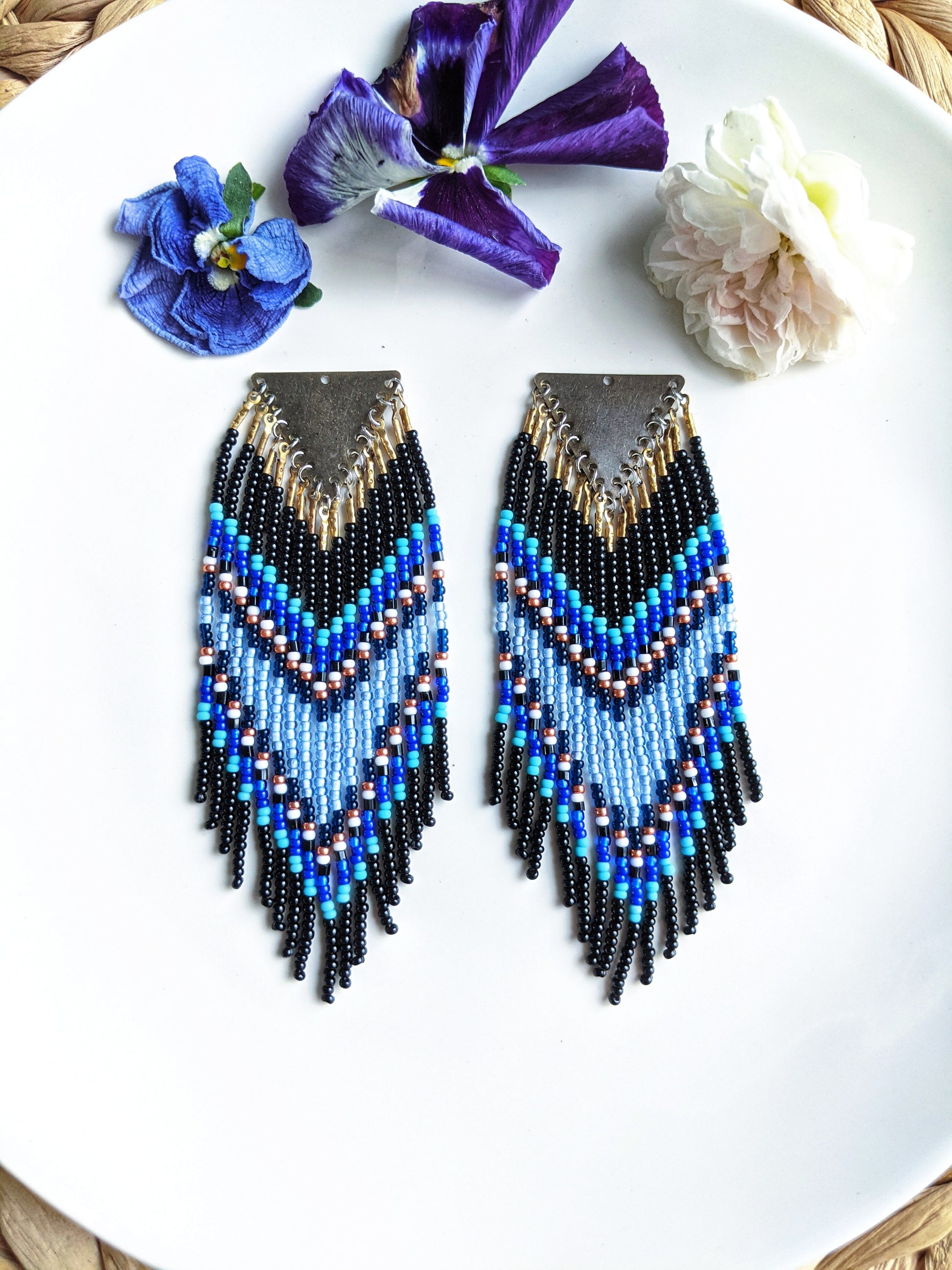Moon & Milk handmade chevron beaded earrings in black, cobalt, and sky blue with metallic accents, styled on a white plate with fresh flowers.