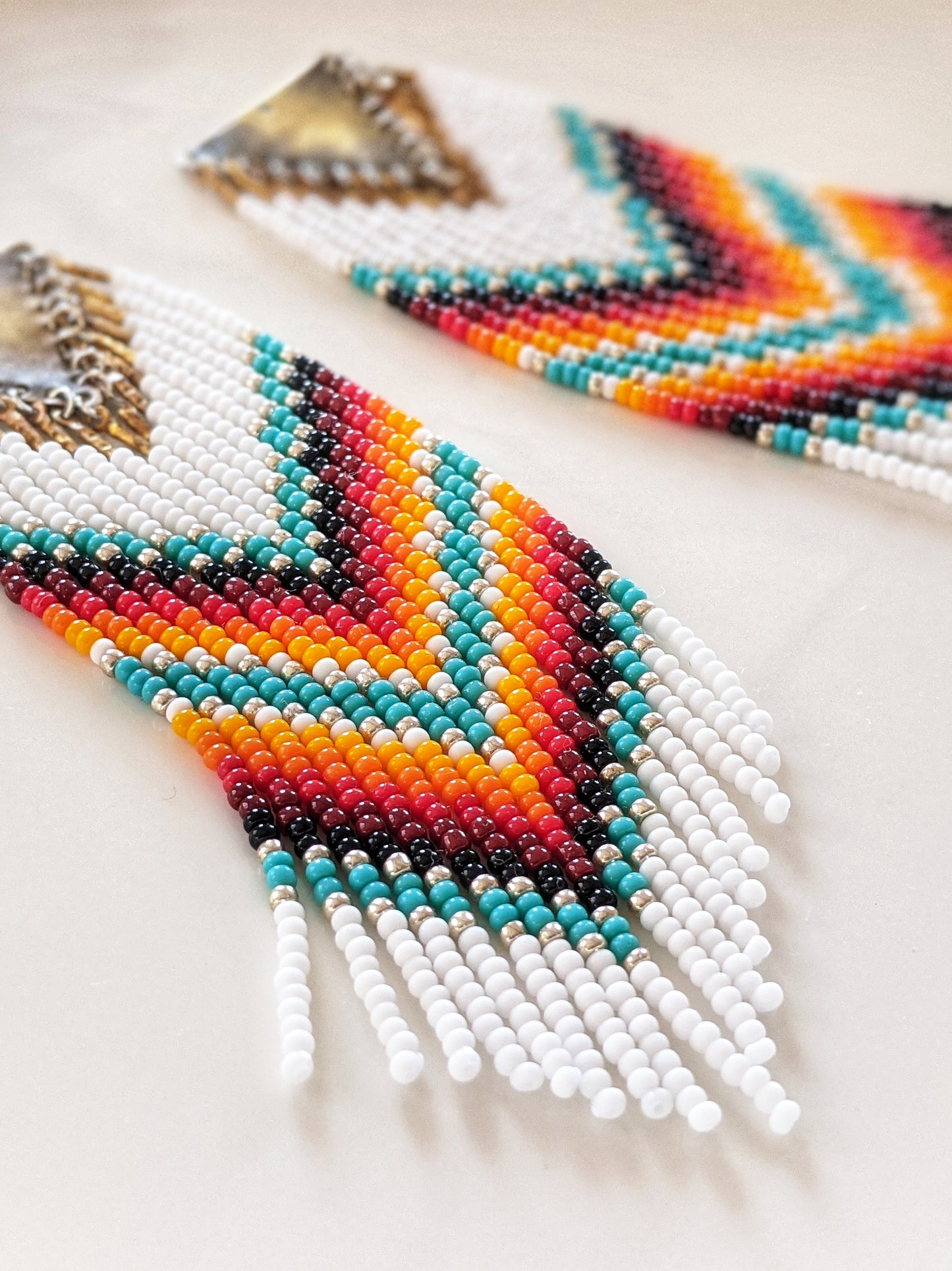 Handwoven chevron earrings by Moon & Milk featuring red, turquoise, yellow, and white seed beads with brass triangle tops, styled on a white ceramic plate.