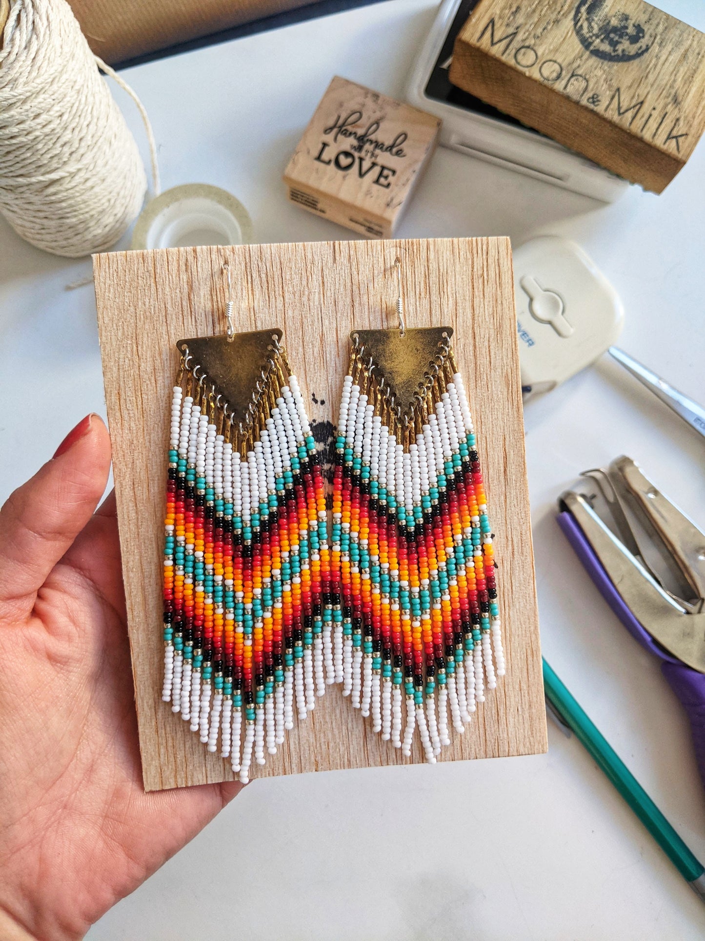 Handwoven chevron earrings by Moon & Milk featuring red, turquoise, yellow, and white seed beads with brass triangle tops, styled on balsa wood.