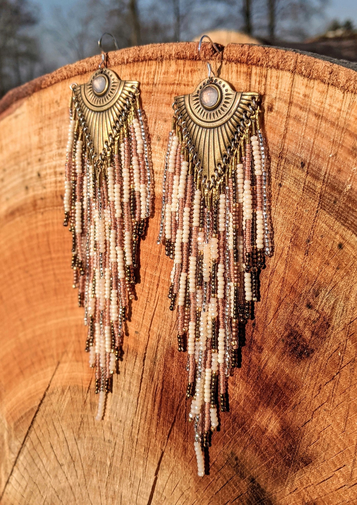 Moon & Milk long beaded earrings with brass fan-shaped medallions and cascading fringes of blush, cream, bronze, and rose gold beads on a tree trunk.