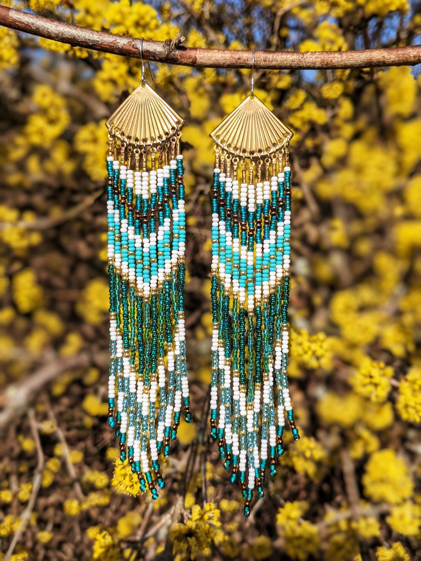 Moon & Milk handcrafted long native earrings with white, turquoise, gold, and deep green seed beads and brass fan tops, styled outdoors in spring.
