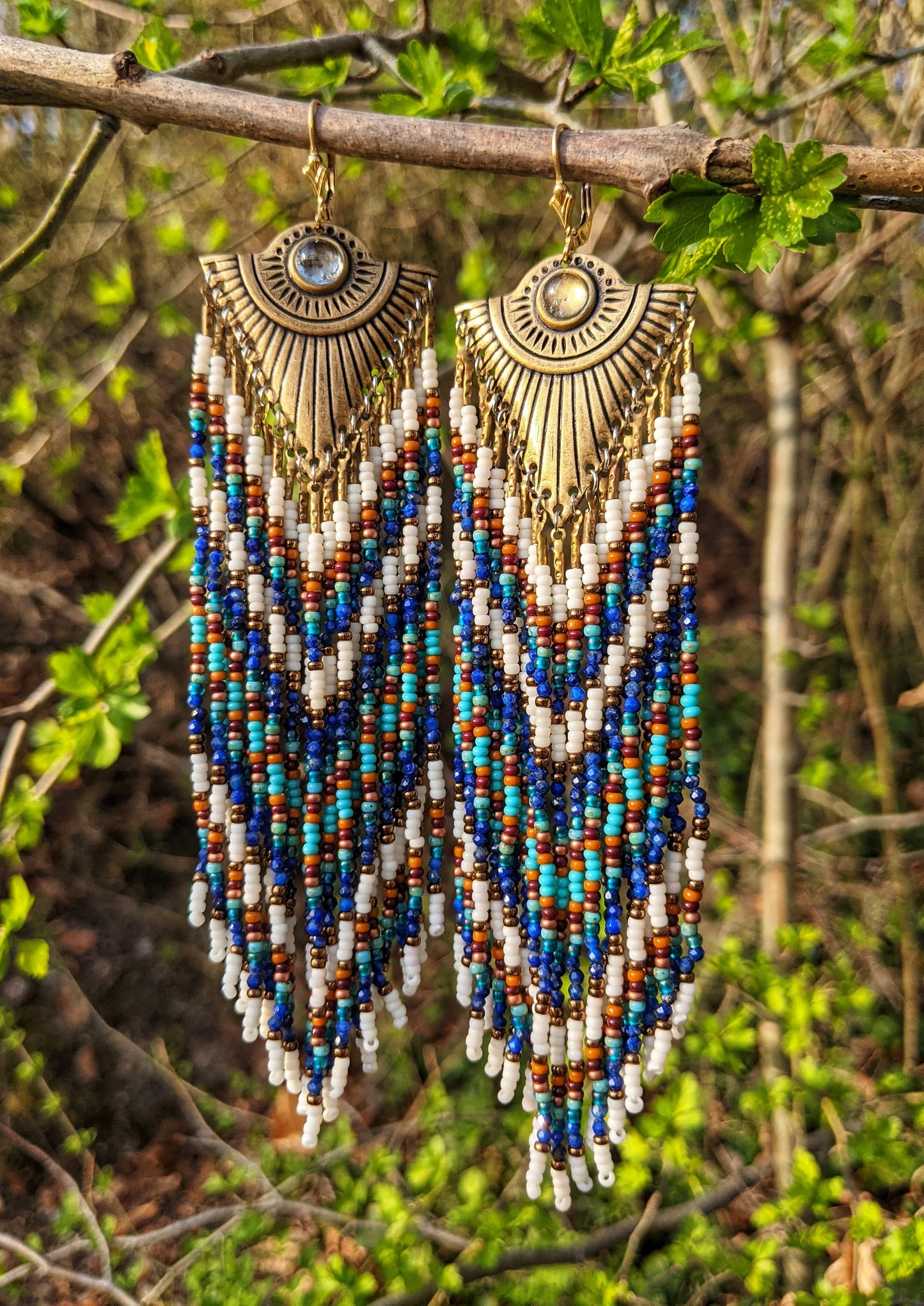 Bohemian-inspired earrings with brass sunburst details, in earthy tones of blue, cream, and bronze, displayed outdoors with green foliage.