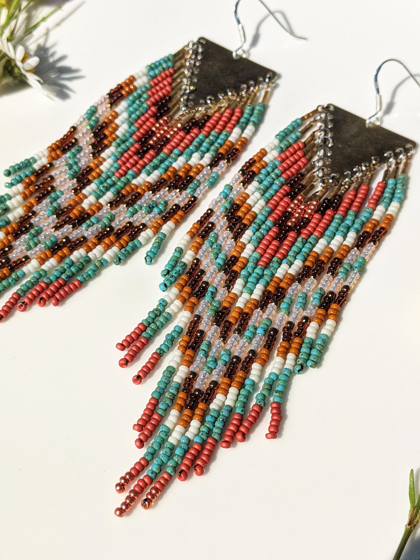 Colorful beaded earrings displayed on a white plate in sunlight, featuring a chevron pattern with shades of teal, coral, and brown, accented by a triangle brass hardware.