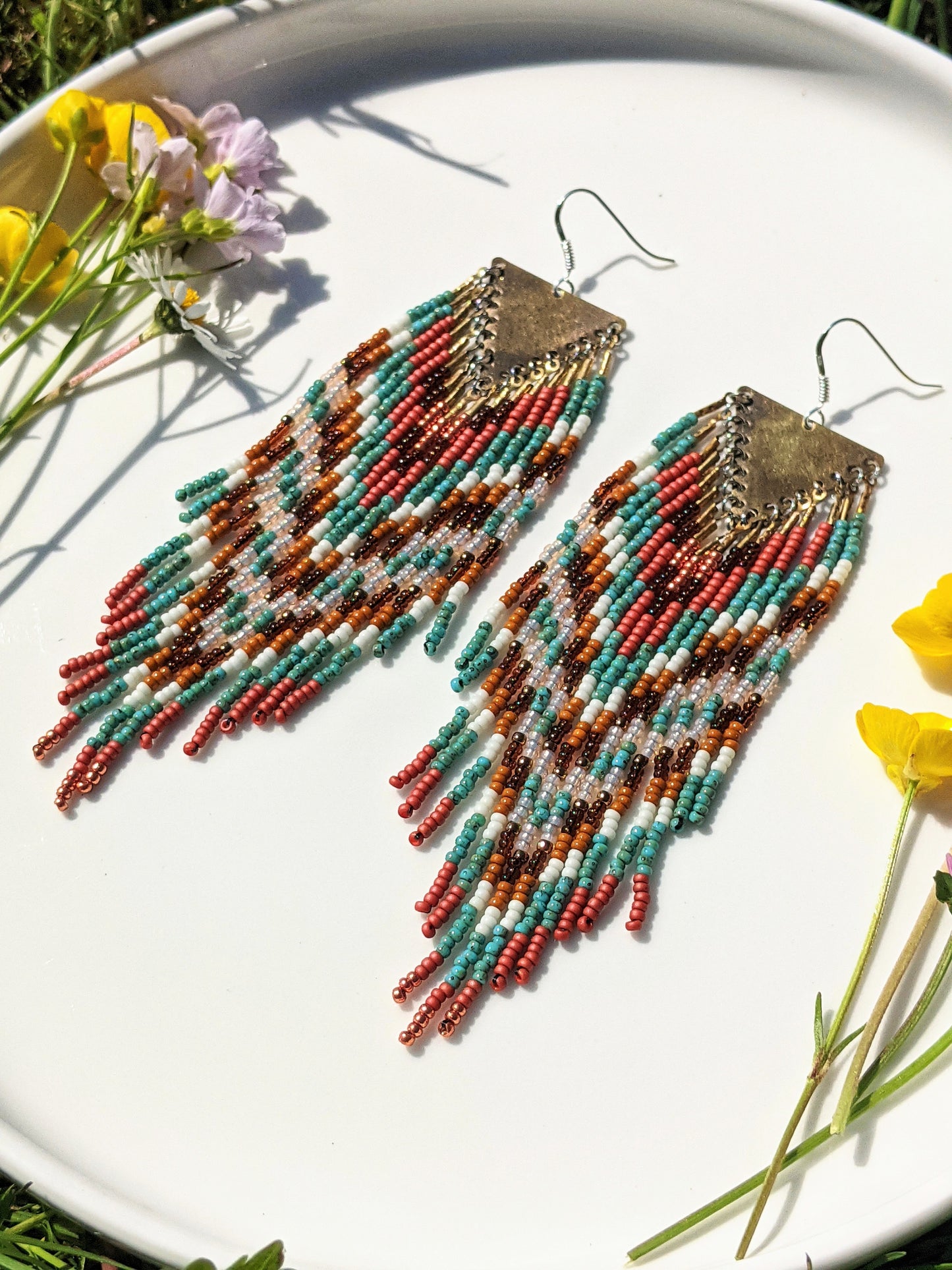 Colorful beaded earrings displayed on a white plate in sunlight, featuring a chevron pattern with shades of teal, coral, and brown, accented by a triangle brass hardware.