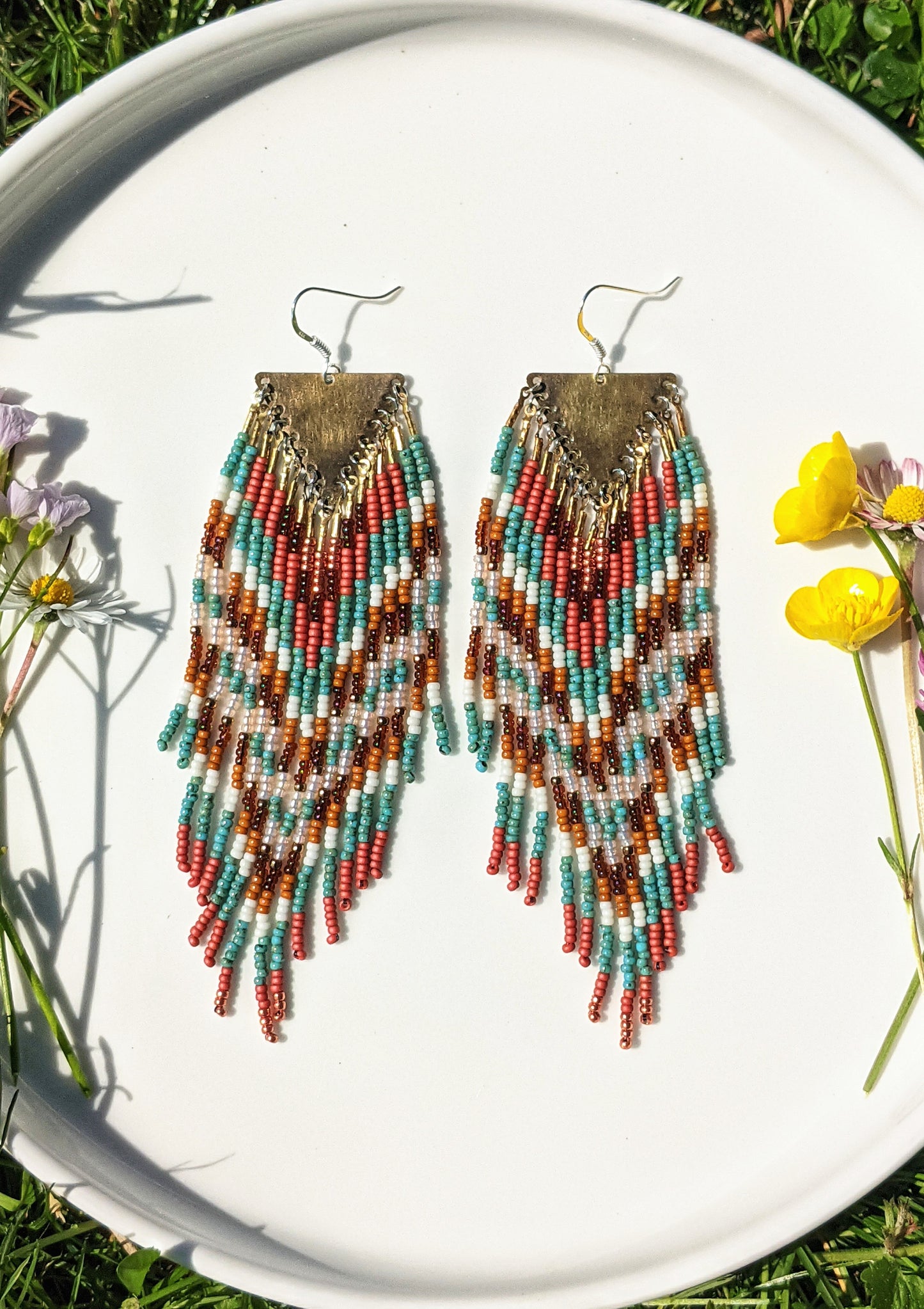 Colorful beaded earrings displayed on a white plate in sunlight, featuring a chevron pattern with shades of teal, coral, and brown, accented by a triangle brass hardware.