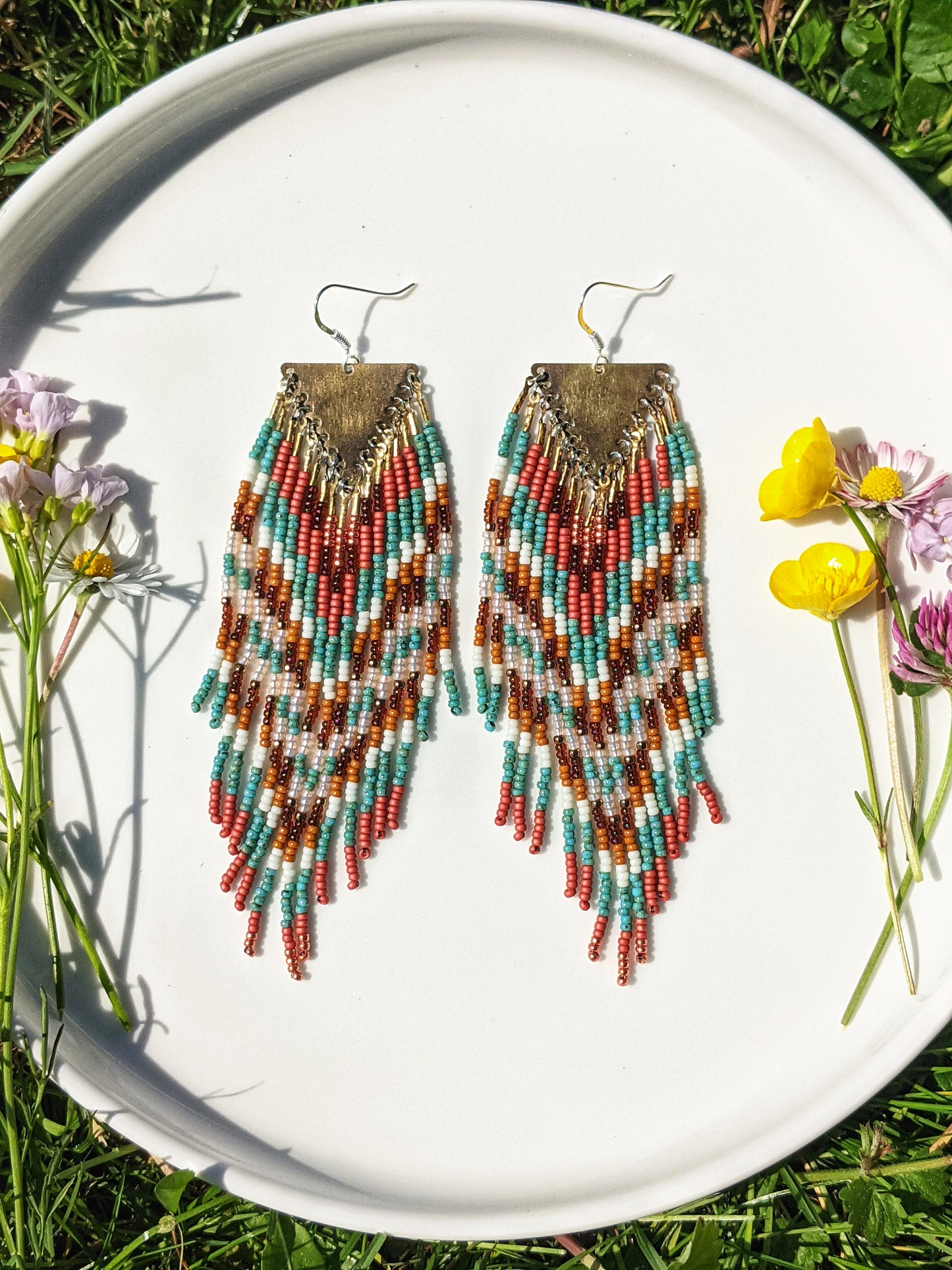 Colorful beaded earrings displayed on a white plate in sunlight, featuring a chevron pattern with shades of teal, coral, and brown, accented by a triangle brass hardware.