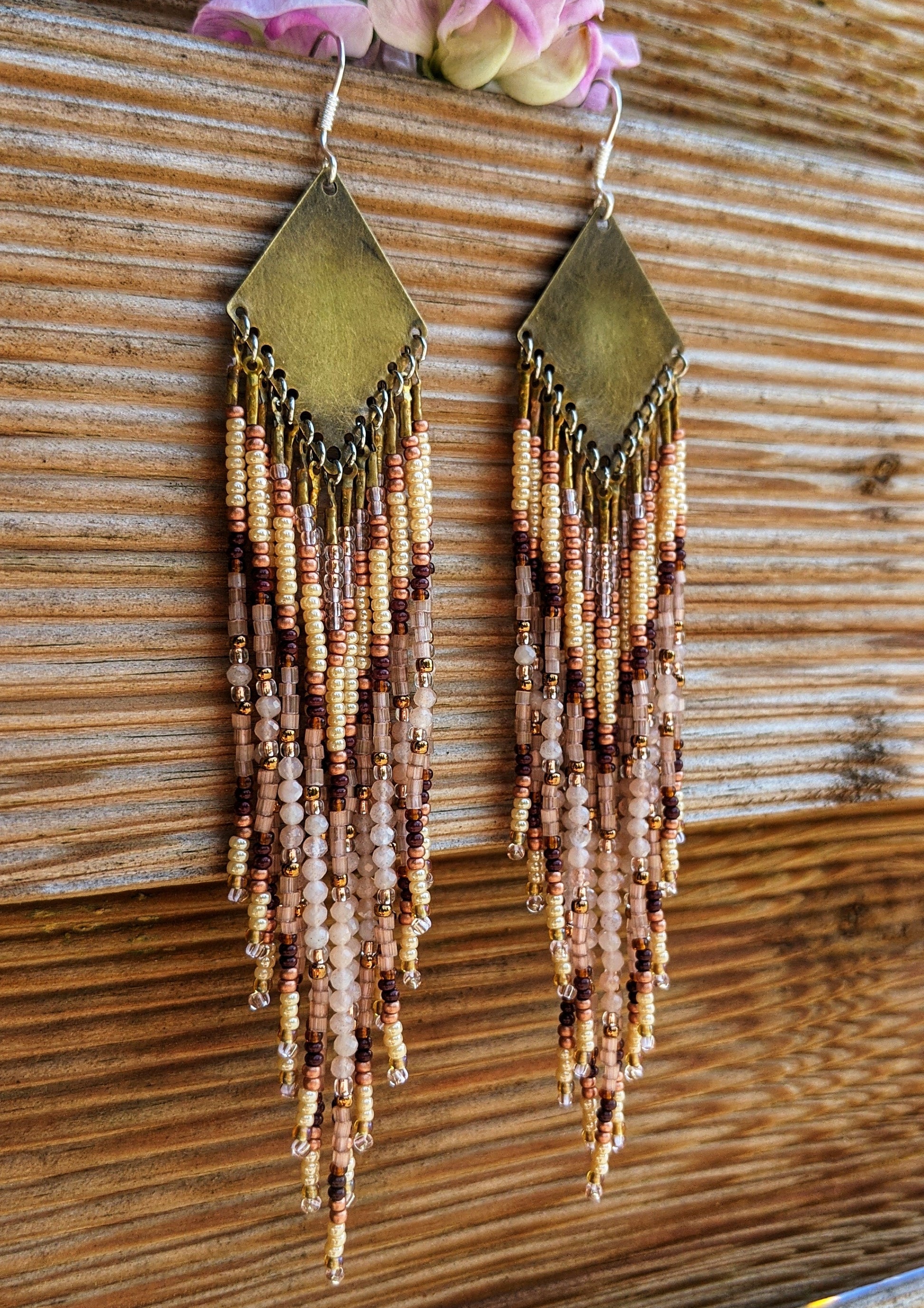 Moon & Milk - Long seed bead earrings with gold, brown, and beige beads in a chevron pattern. Gold diamond-shaped tops. Photographed on a wooden background with pink flowers.