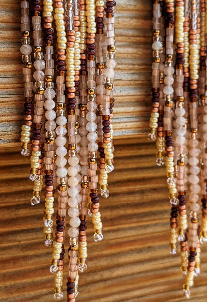 Moon & Milk - Long seed bead earrings with gold, brown, and beige beads in a chevron pattern. Gold diamond-shaped tops. Photographed on a wooden background with pink flowers.