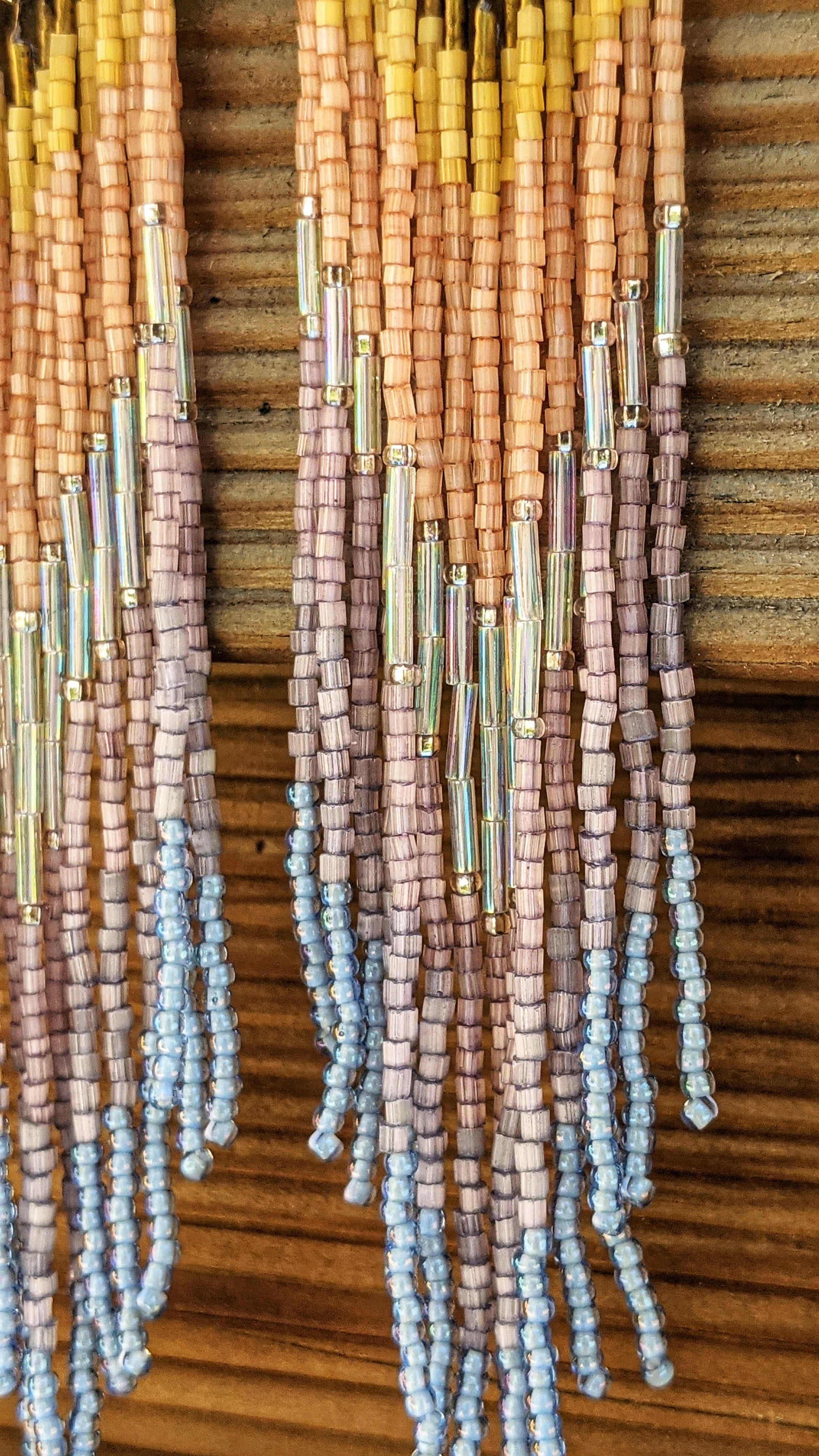 Handcrafted beaded earrings with a soft ombre fringe design, transitioning from yellow to peach to lavender and ending in light blue. The earrings feature a brass top for a colorful bohemian look.