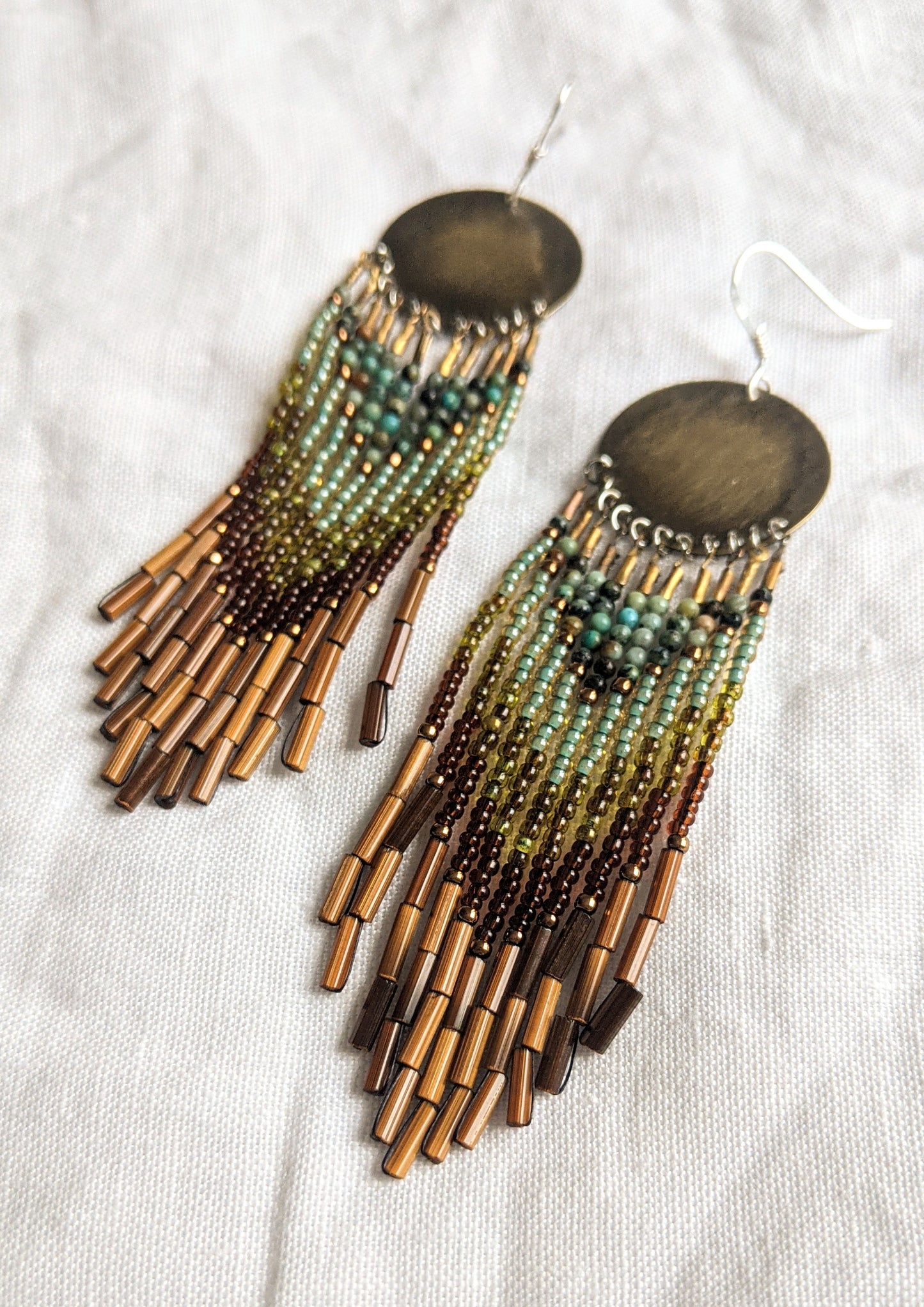 Moon & Milk handcrafted earrings with round brass tops, green and brown beadwork, and layered bamboo bugle fringe displayed on a soft white fabric background.