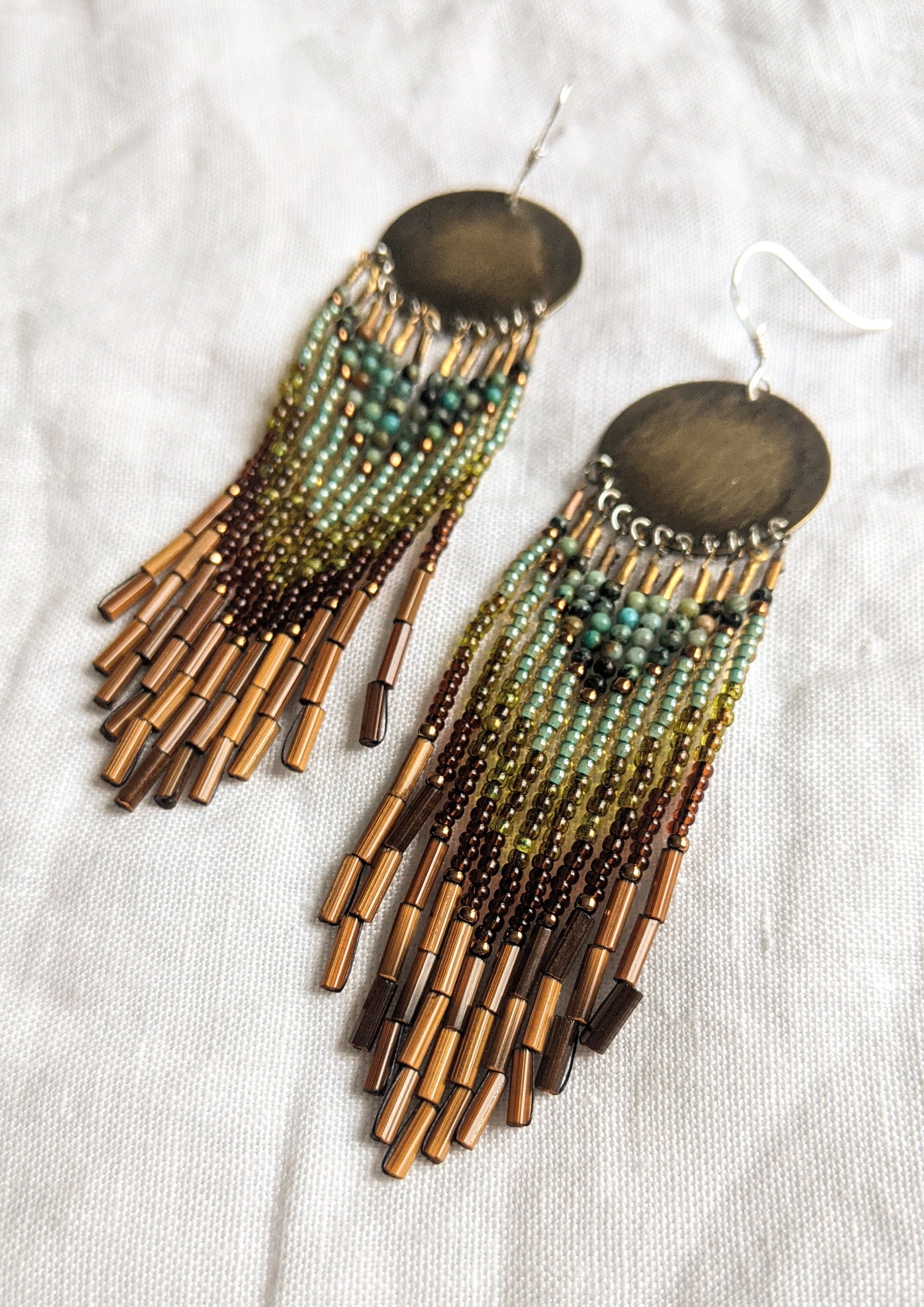Moon & Milk handcrafted earrings with round brass tops, green and brown beadwork, and layered bamboo bugle fringe displayed on a soft white fabric background.