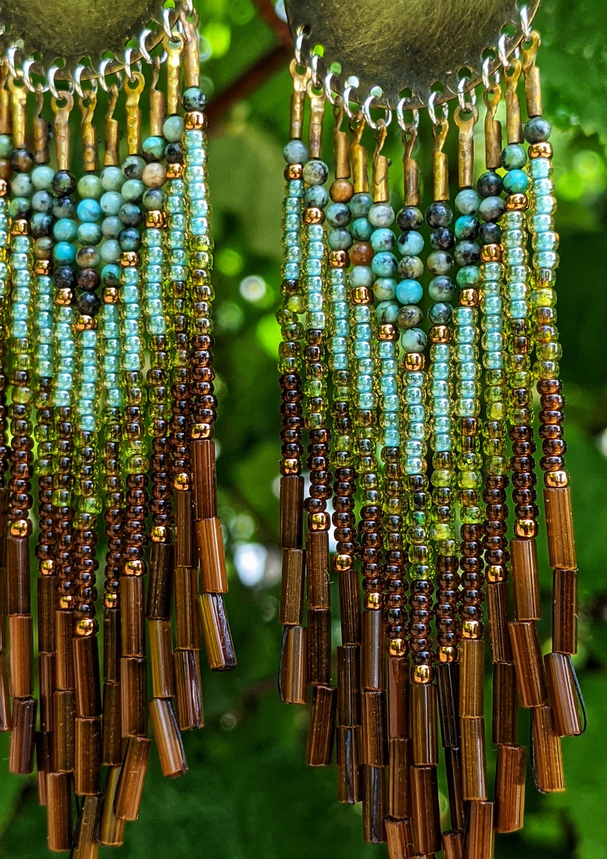 Moon & Milk artisan earrings with brass discs, green and turquoise beadwork, earthy brown accents, and long bugle fringe hanging in front of green leaves.
