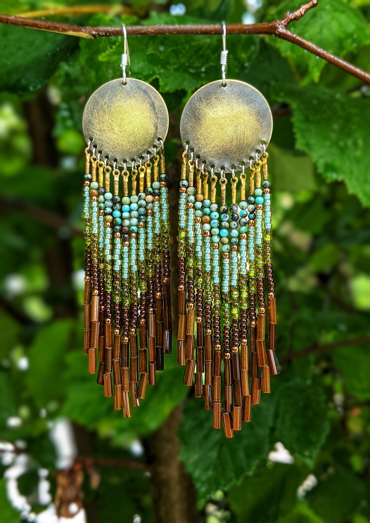 Moon & Milk artisan earrings with brass discs, green and turquoise beadwork, earthy brown accents, and long bugle fringe hanging in front of green leaves.