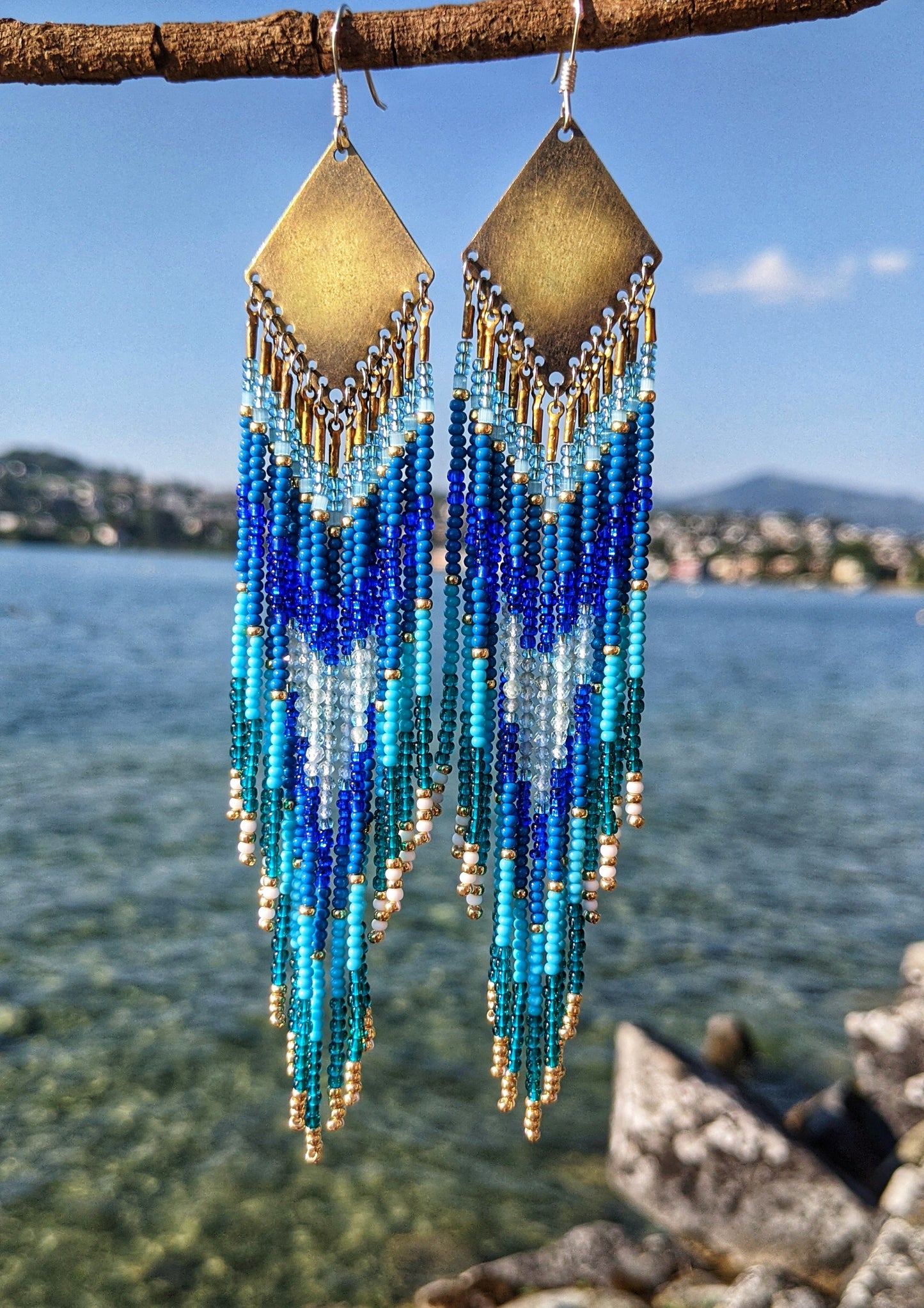Moon & Milk handcrafted earrings with turquoise, cobalt, and gold seed beads, inspired by Swiss lakes and alpine clarity, hanging against a blue lake.