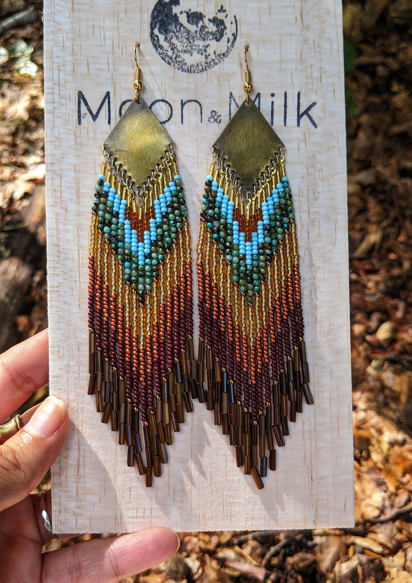 Moon & Milk - Long boho beaded earrings with gold, turquoise, brown, and maroon beads in a chevron pattern. Gold brass diamond-shaped tops. Displayed on a "Moon & Milk" branded card.