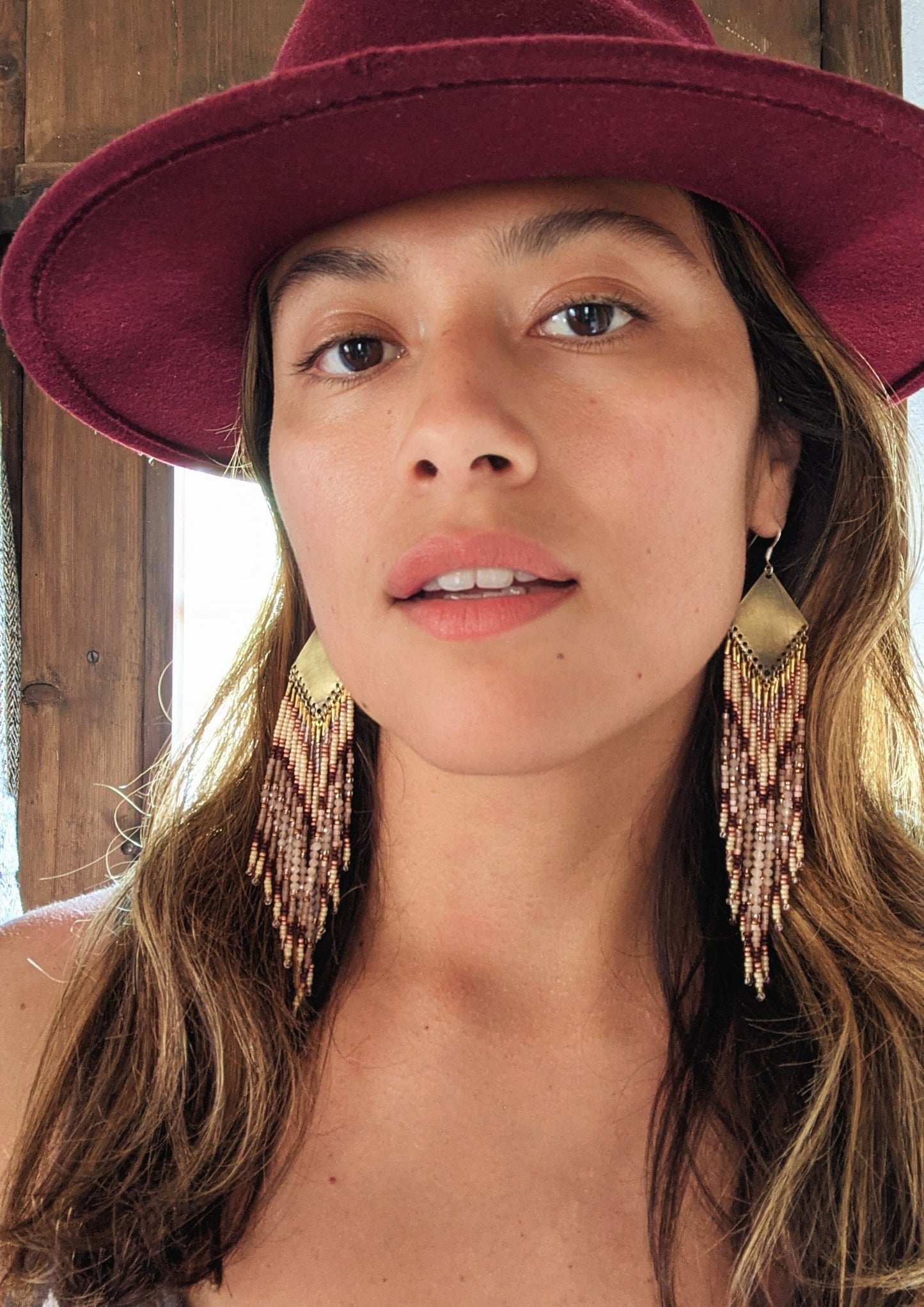 Moon & Milk - Woman wearing long beaded earrings with gold, brown, and beige beads in a chevron pattern. She is also wearing a burgundy hat.
