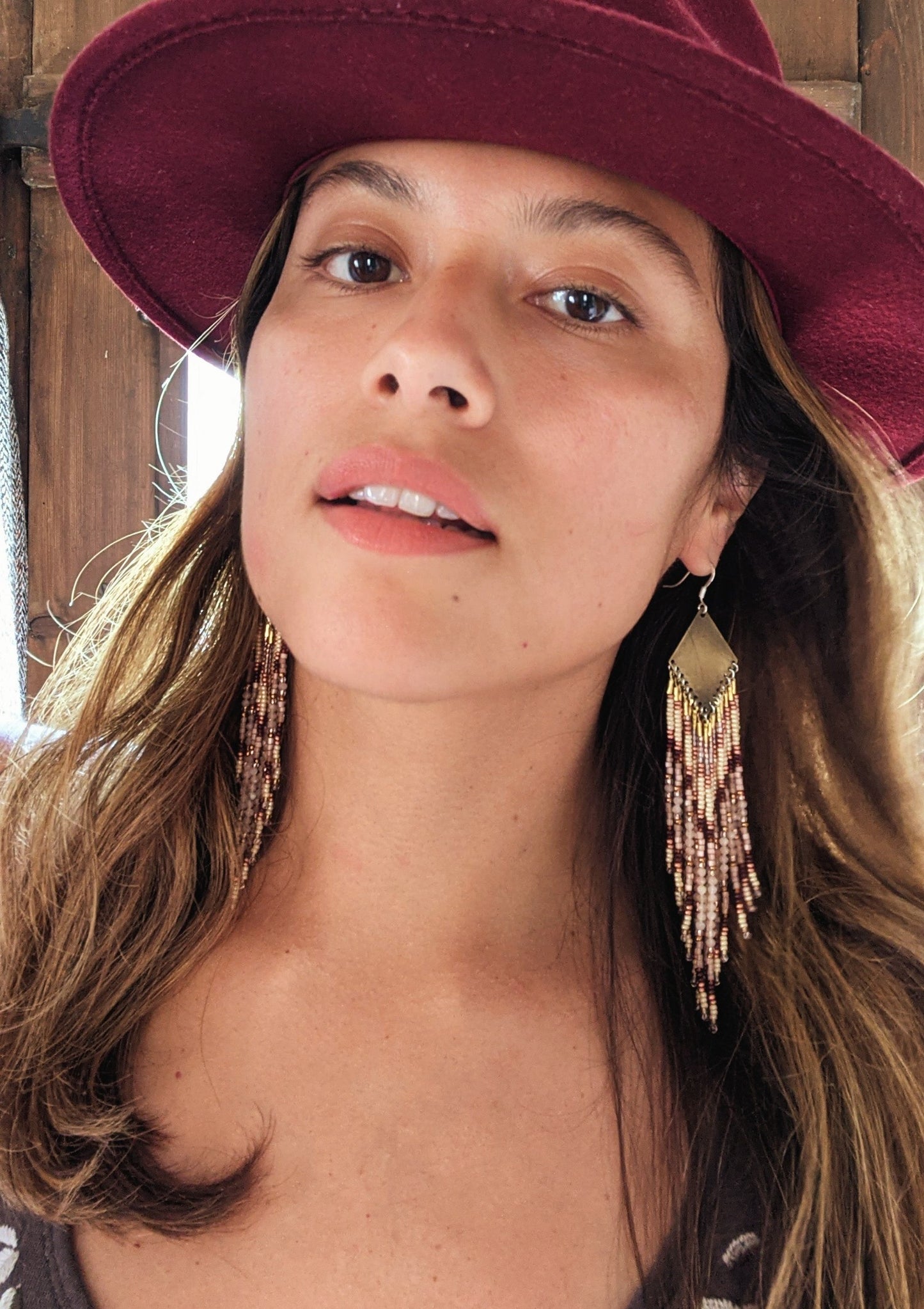 Moon & Milk - Woman wearing long beaded earrings with gold, brown, and beige beads in a chevron pattern. She is also wearing a burgundy hat.