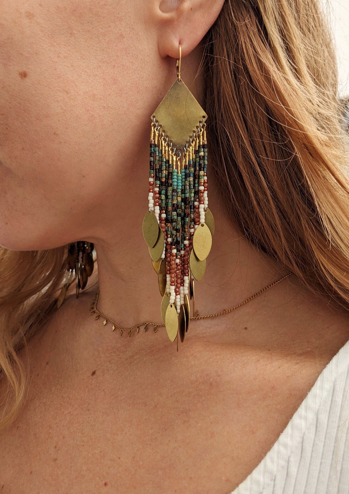 A person wearing a gold-colored beaded earring with intricate geometric patterns, made with antique-finish brass and glass beads.