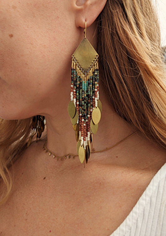 A person wearing a gold-colored beaded earring with intricate geometric patterns, made with antique-finish brass and glass beads.