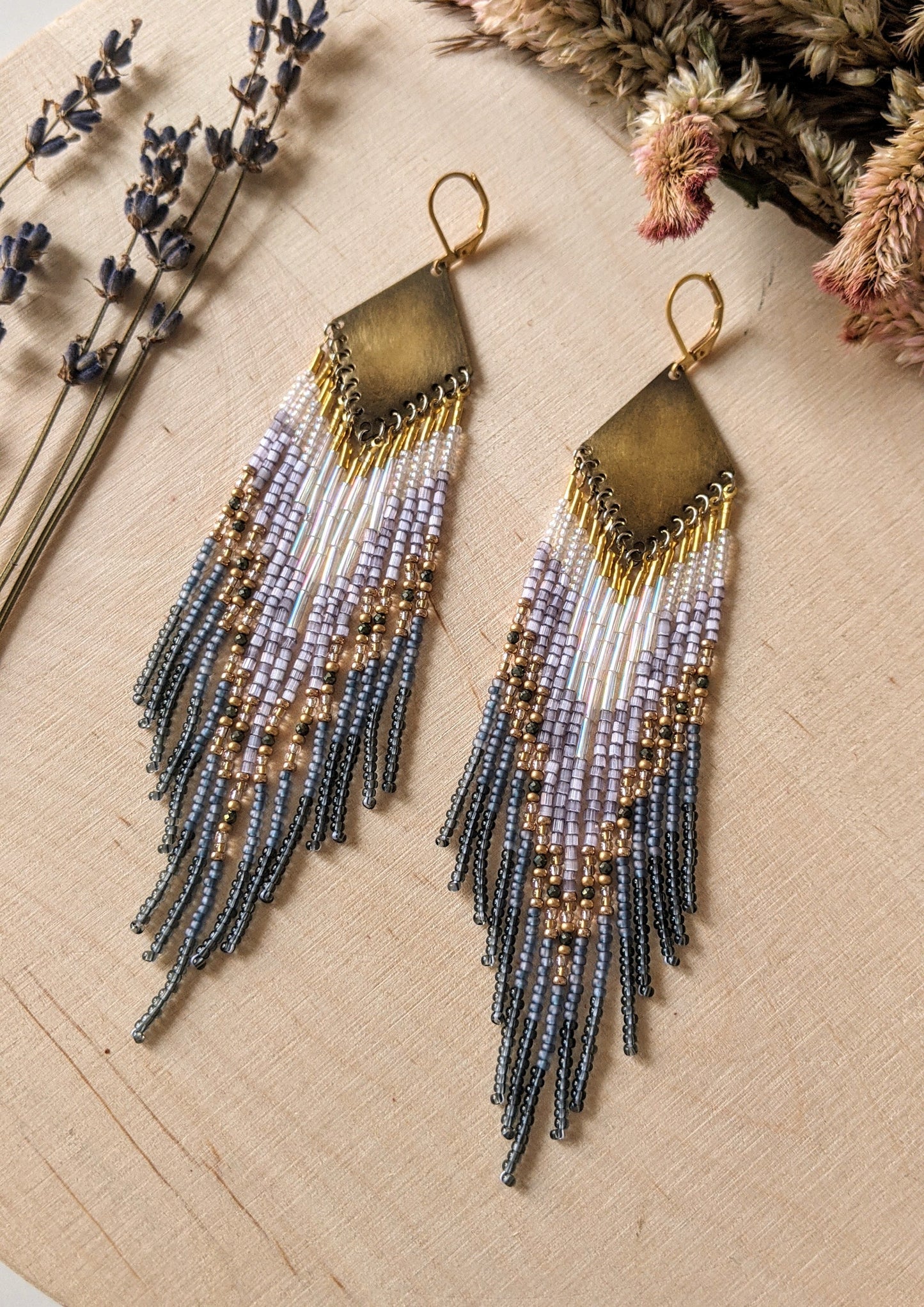 Moon & Milk handcrafted beaded earrings with lavender, gold, grey, and iridescent seed beads on brass diamond-shaped findings, displayed on a wood background.