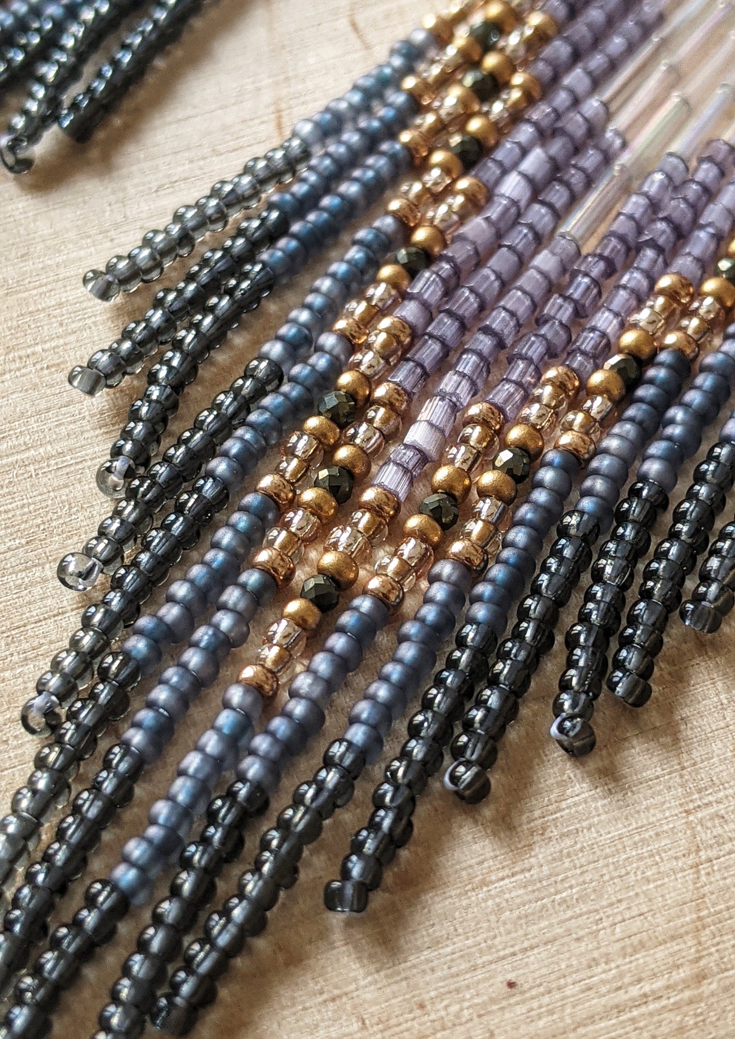 Close-up of handcrafted beaded earrings with lavender, gold, grey, and iridescent seed beads on brass diamond-shaped findings, displayed on a wood background.