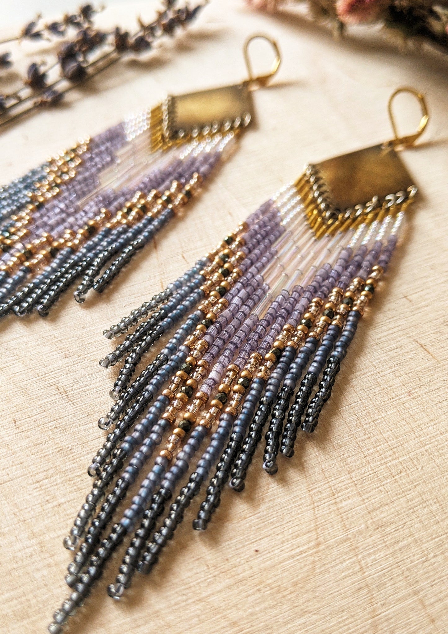 Moon & Milk handcrafted beaded earrings with lavender, gold, grey, and iridescent seed beads on brass diamond-shaped findings, displayed on a wood background.