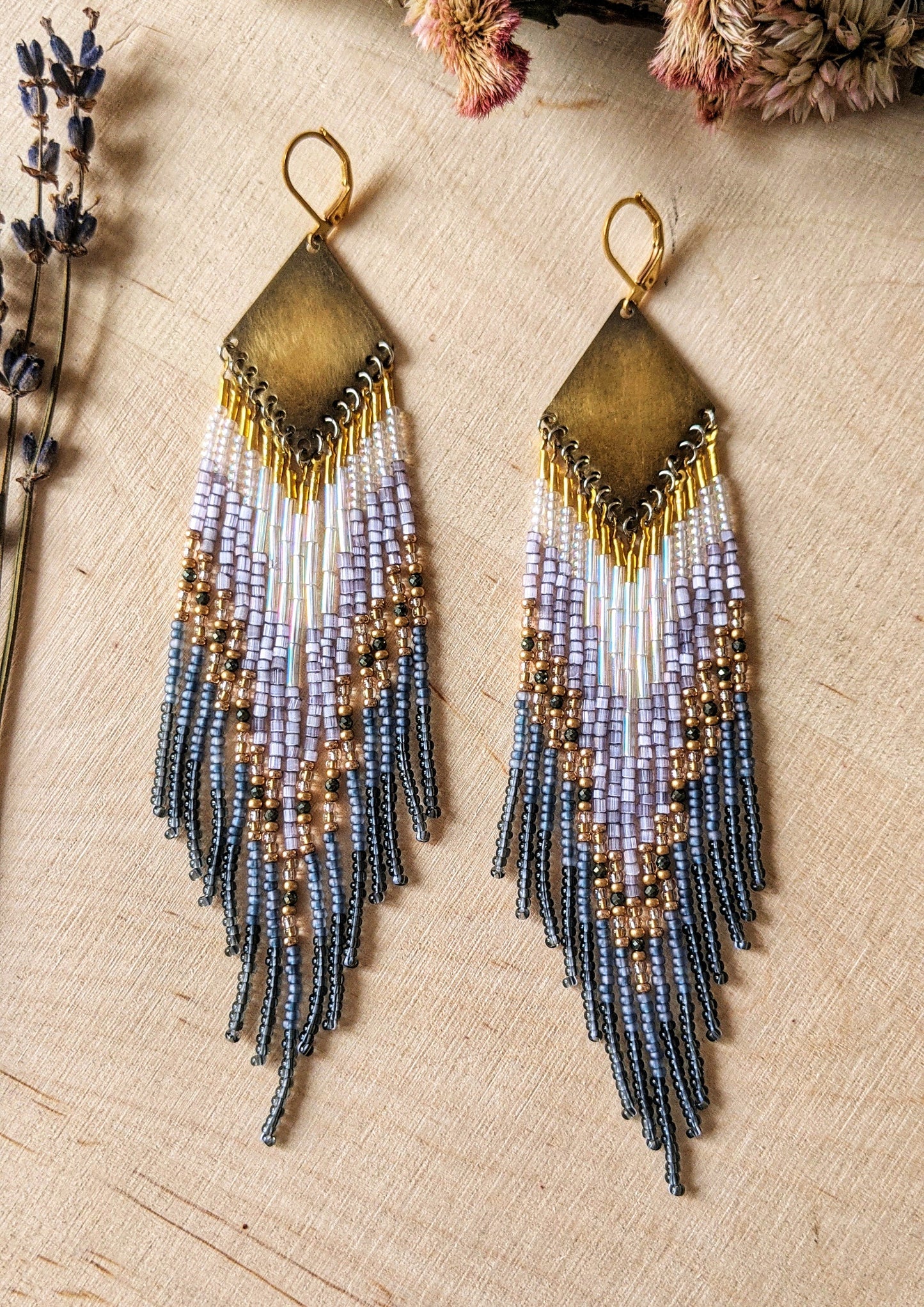 Moon & Milk handcrafted beaded earrings with lavender, gold, grey, and iridescent seed beads on brass diamond-shaped findings, displayed on a wood background.
