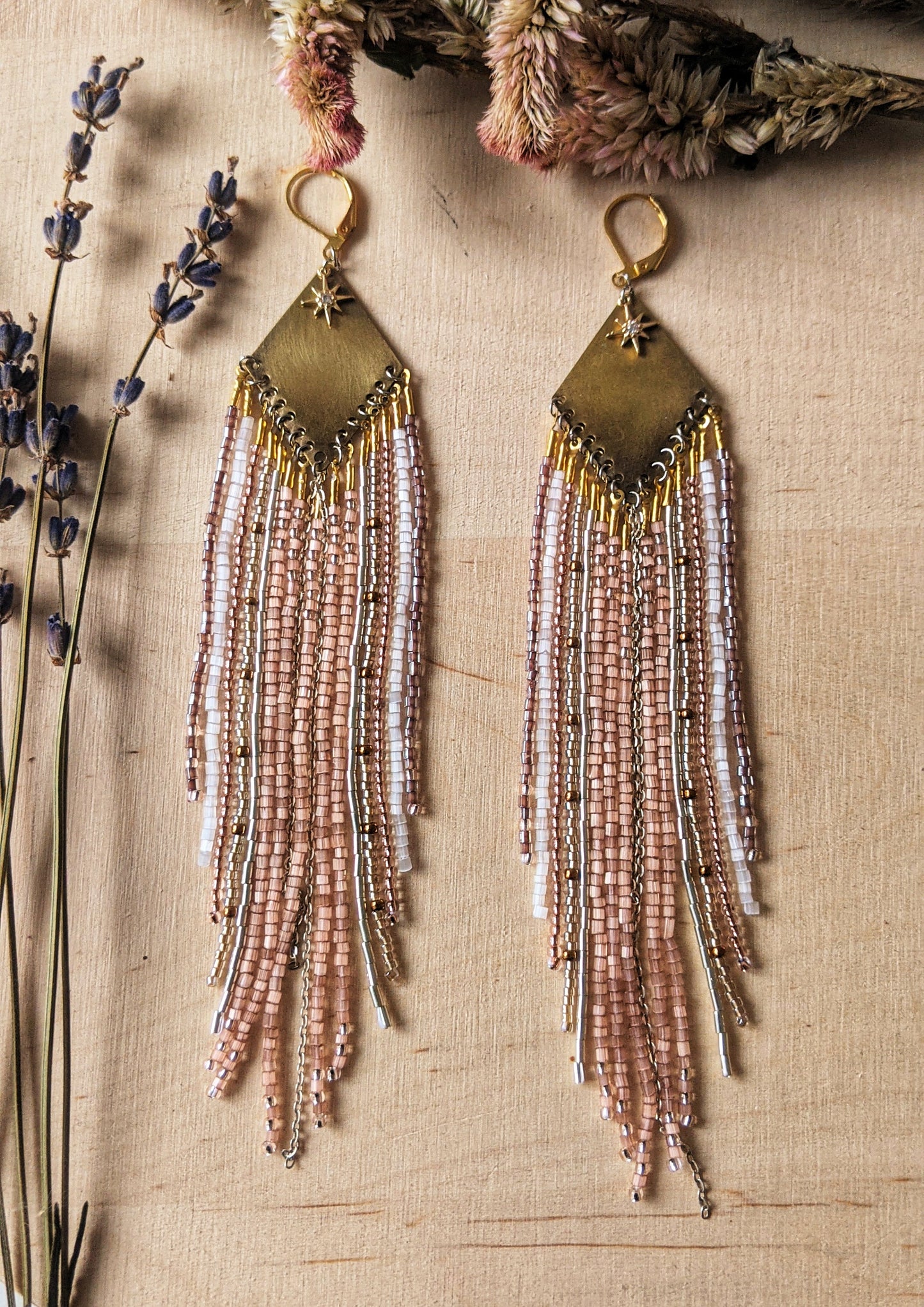 Moon & Milk earrings with a brass diamond top and soft blush, white, and metallic fringe beadwork, photographed on a wood surface with dried florals.