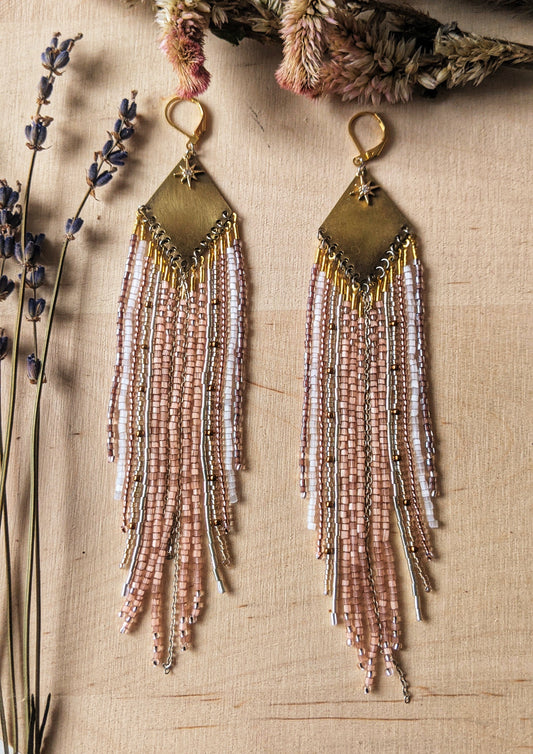 Moon & Milk earrings with a brass diamond top and soft blush, white, and metallic fringe beadwork, photographed on a wood surface with dried florals.