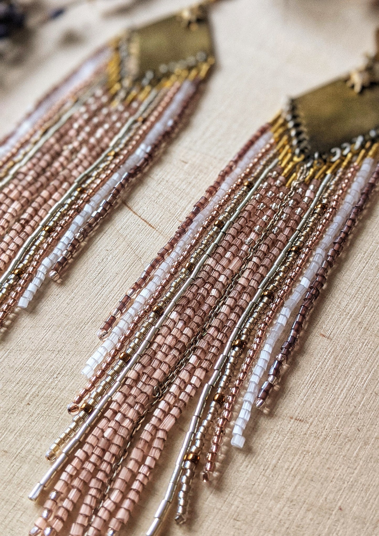 Moon & Milk earrings with a brass diamond top and soft blush, white, and metallic fringe beadwork, photographed on a wood surface with dried florals.
