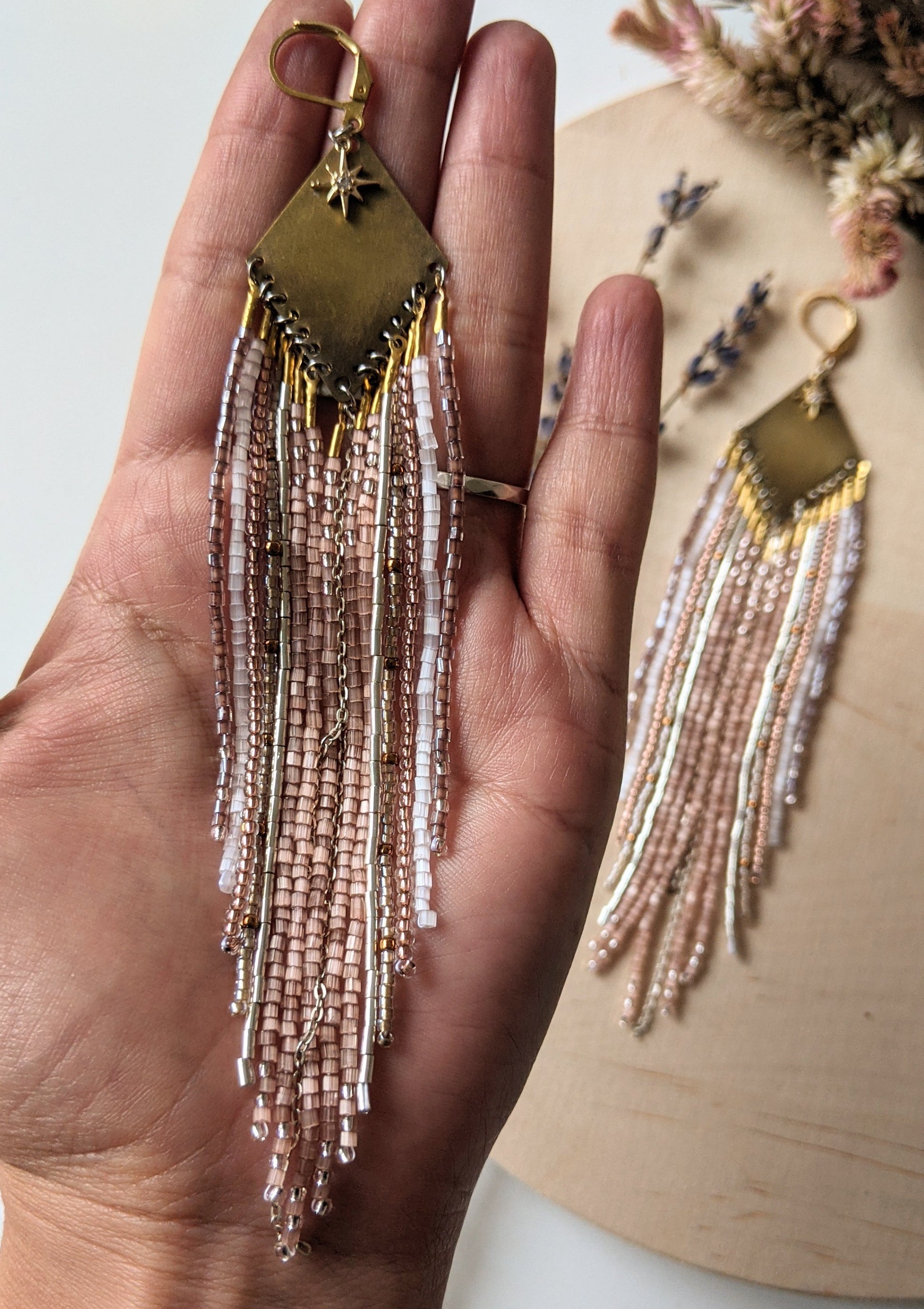 Moon & Milk earrings with a brass diamond top and soft blush, white, and metallic fringe beadwork, photographed on a wood surface with dried florals.