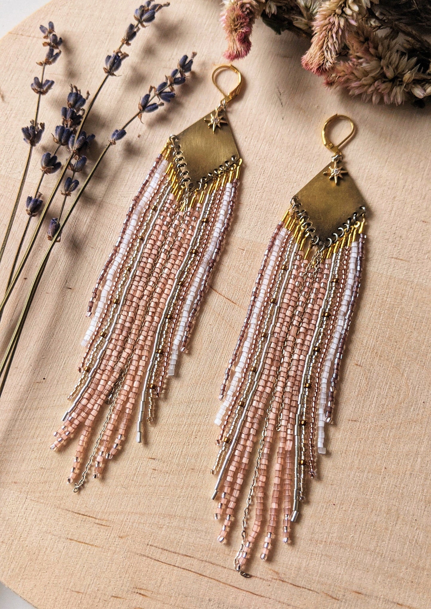 Moon & Milk earrings with a brass diamond top and soft blush, white, and metallic fringe beadwork, photographed on a wood surface with dried florals.
