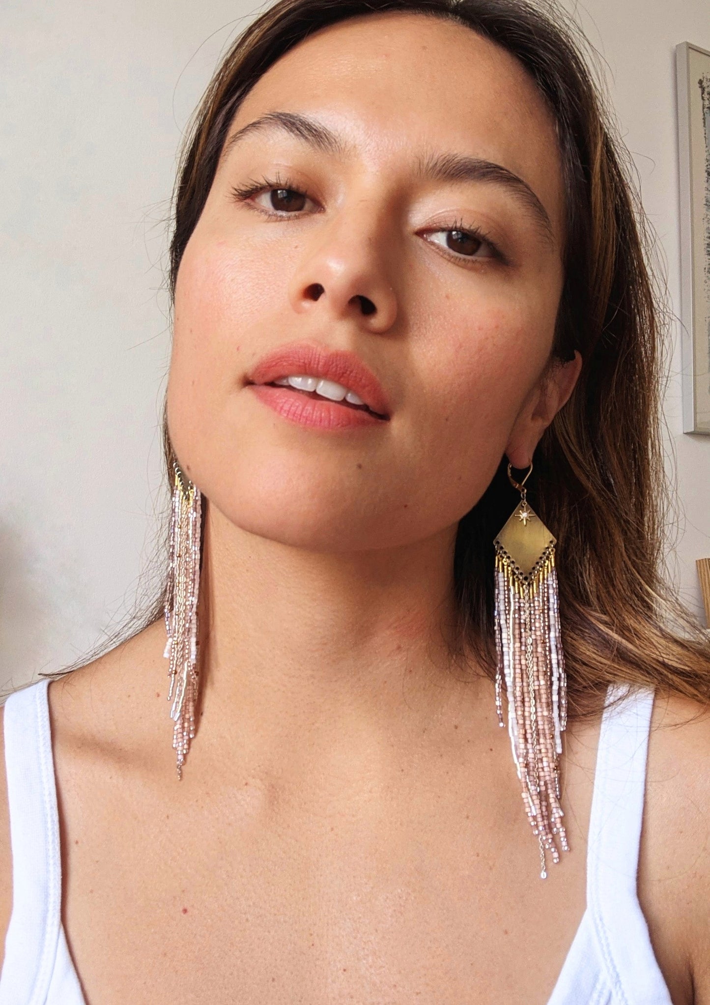 Moon & Milk earrings with a brass diamond top and soft blush, white, and metallic fringe beadwork, worn by a woman.