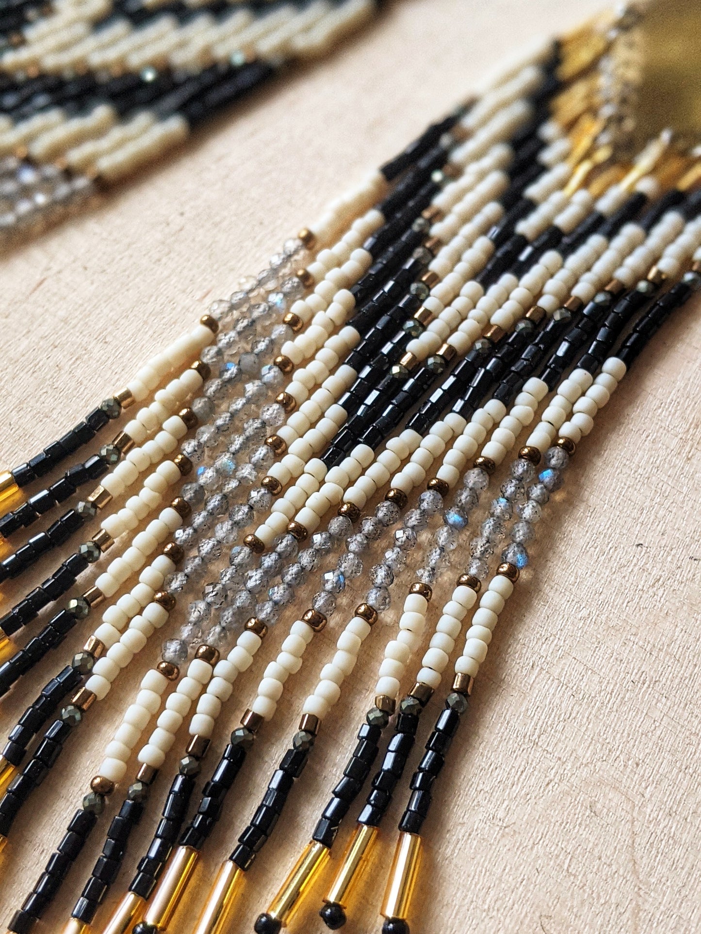 Long seed bead earrings with black, white, and gold beads in a chevron pattern. Gold diamond-shaped tops. Photographed on a light wooden surface.