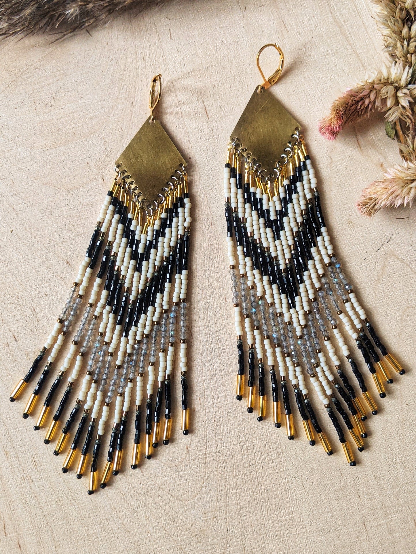 Long beaded earrings with black, white, and gold beads in a chevron pattern. Gold diamond-shaped tops. Photographed on a light wooden surface with dried flowers.- Moon & Milk