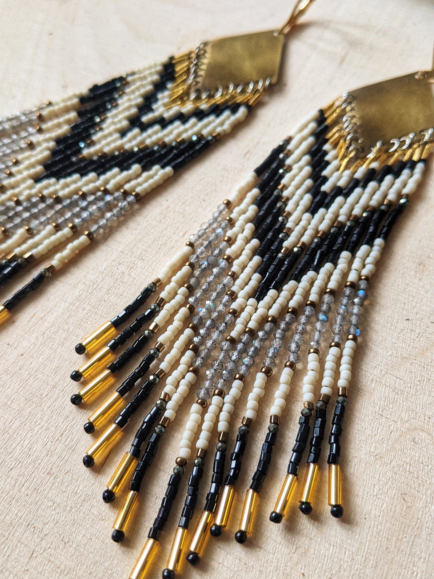 Long seed bead earrings with black, white, and gold beads in a chevron pattern. Gold diamond-shaped tops. Photographed on a light wooden surface with dried flowers.- Moon & Milk