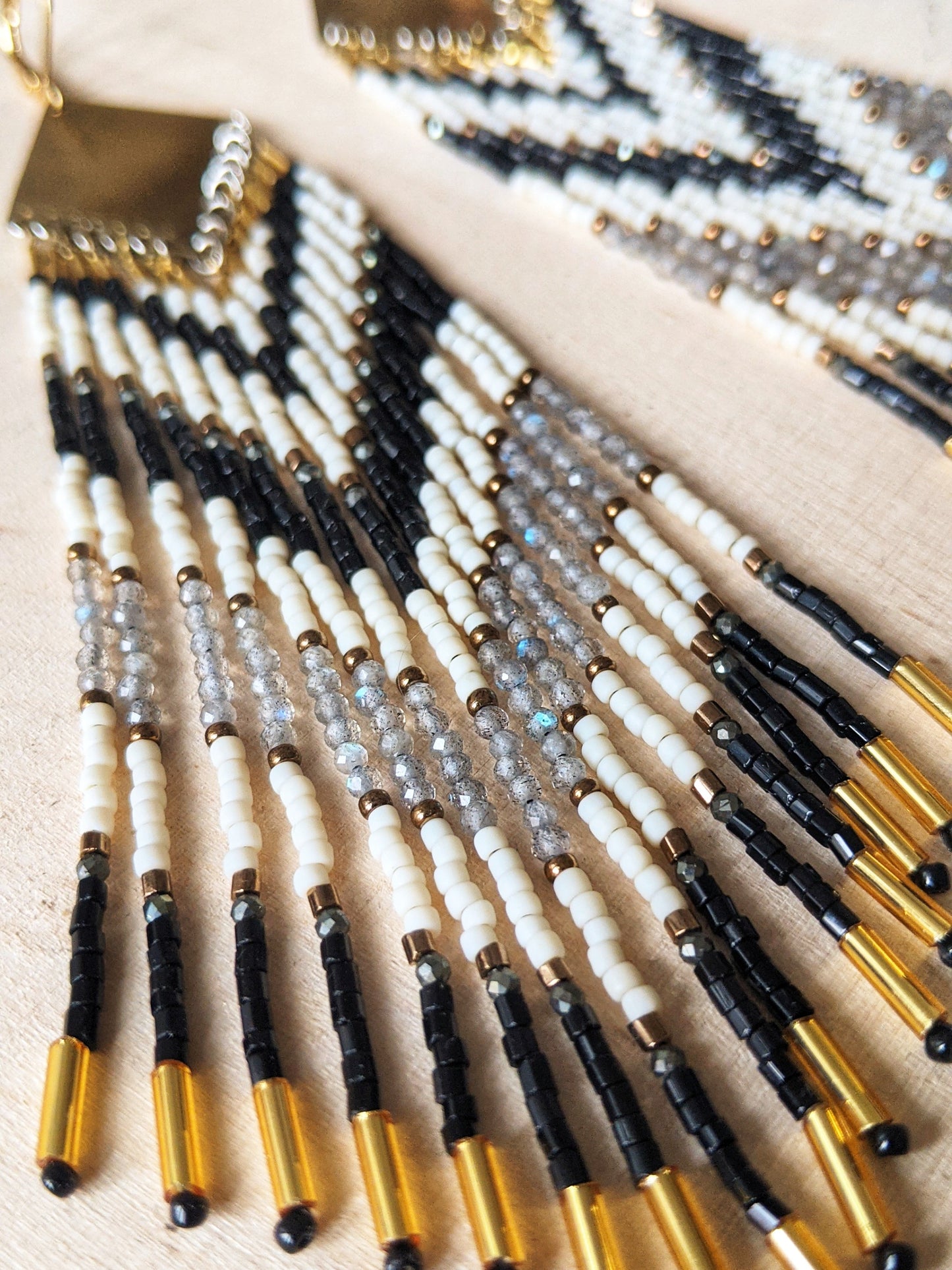 Long seed bead earrings with black, white, and gold beads in a chevron pattern. Gold diamond-shaped tops. Photographed on a light wooden surface.