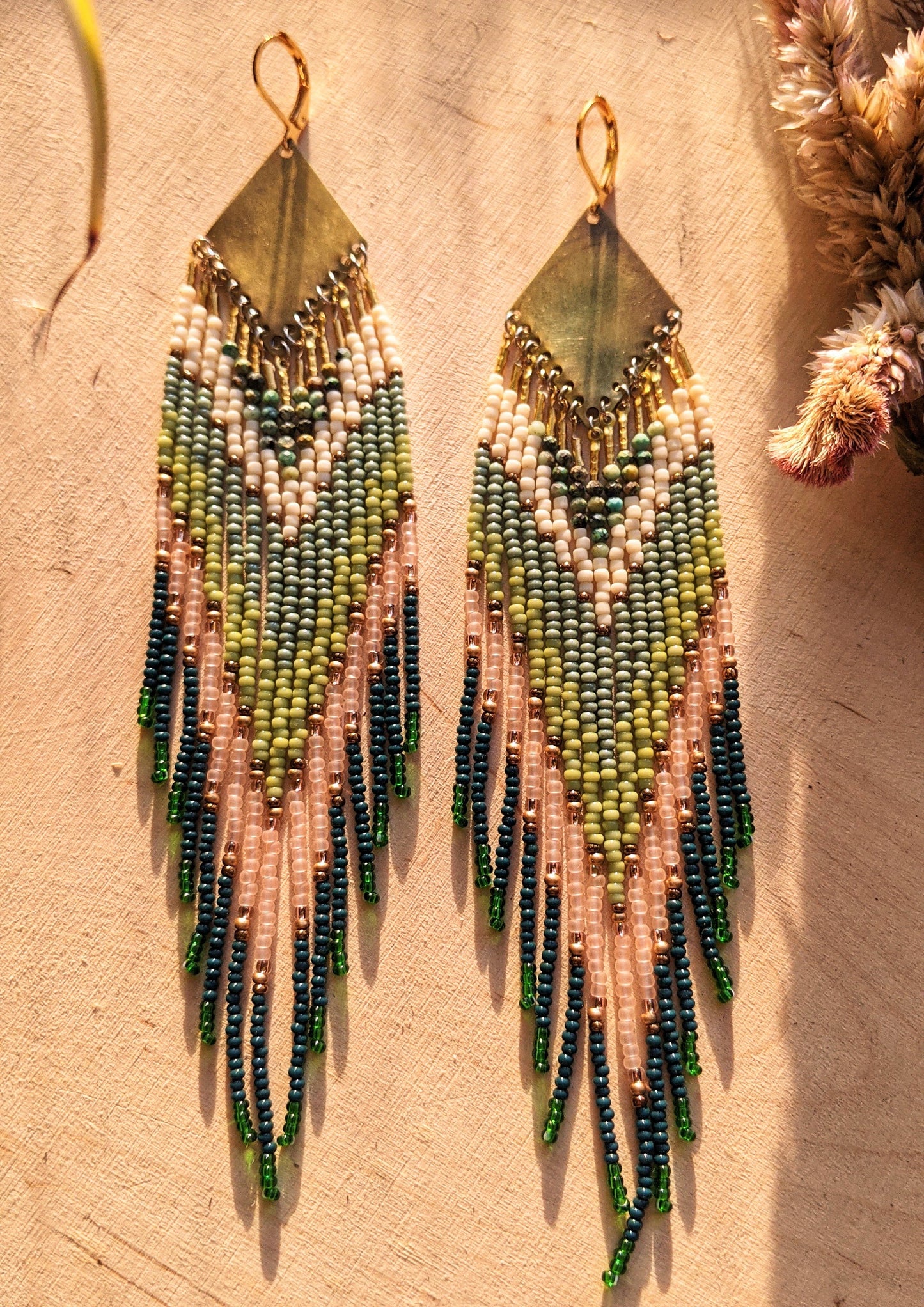Moon & Milk artisan earrings with layered green, blush, cream, and brass beadwork in a chevron pattern, photographed on textured fabric in warm light.