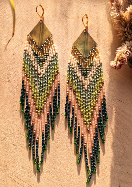 Moon & Milk artisan earrings with layered green, blush, cream, and brass beadwork in a chevron pattern, photographed on textured fabric in warm light.