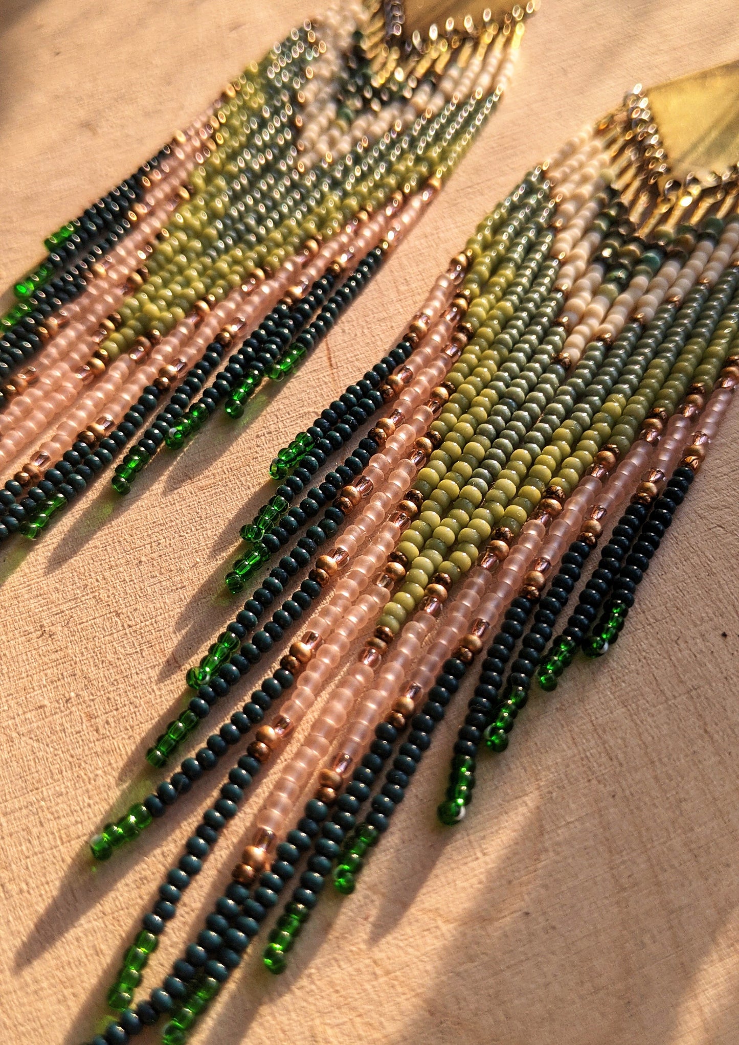 Moon & Milk artisan earrings with layered green, blush, cream, and brass beadwork in a chevron pattern, photographed on textured fabric in warm light.
