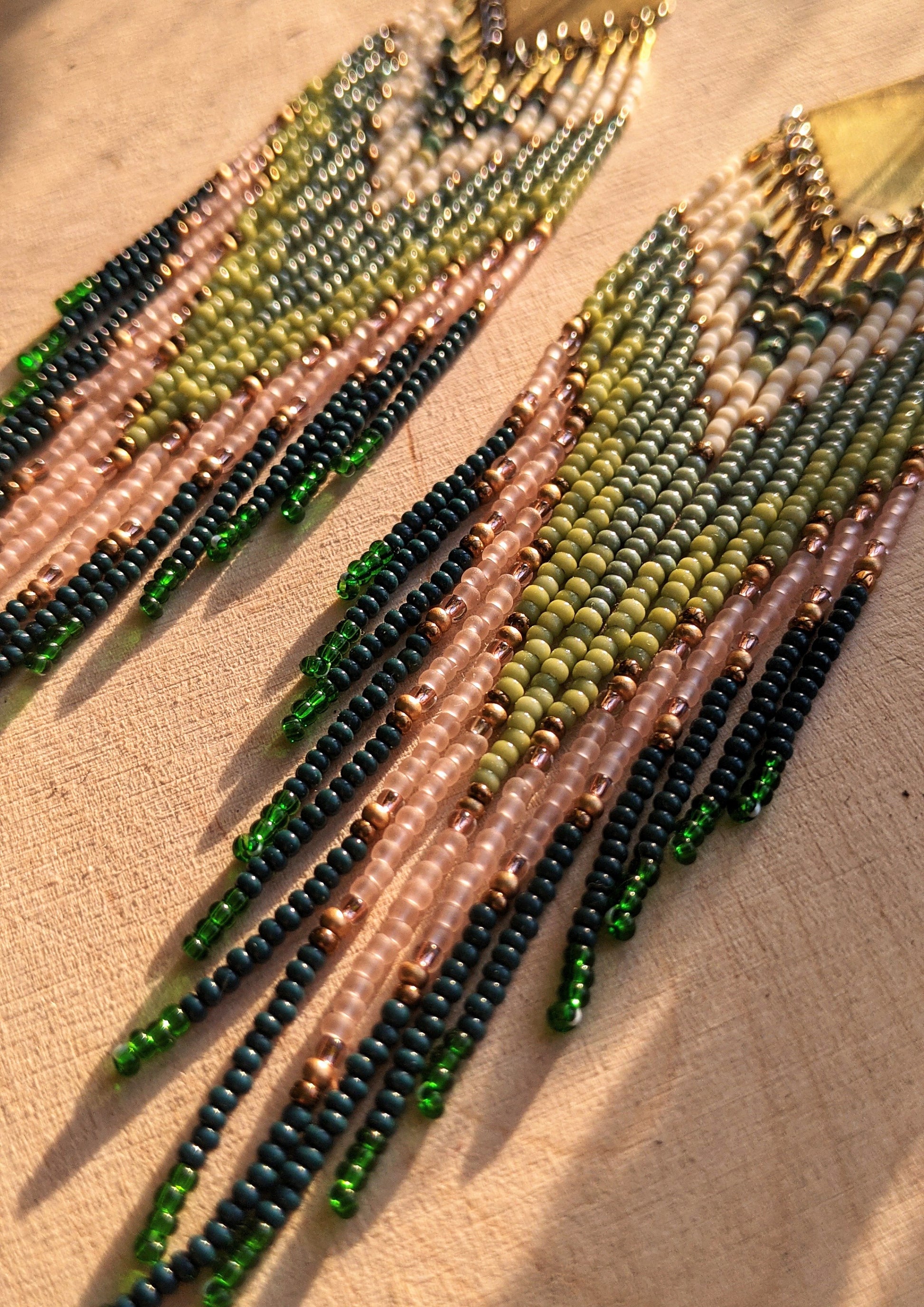 Moon & Milk artisan earrings with layered green, blush, cream, and brass beadwork in a chevron pattern, photographed on textured fabric in warm light.