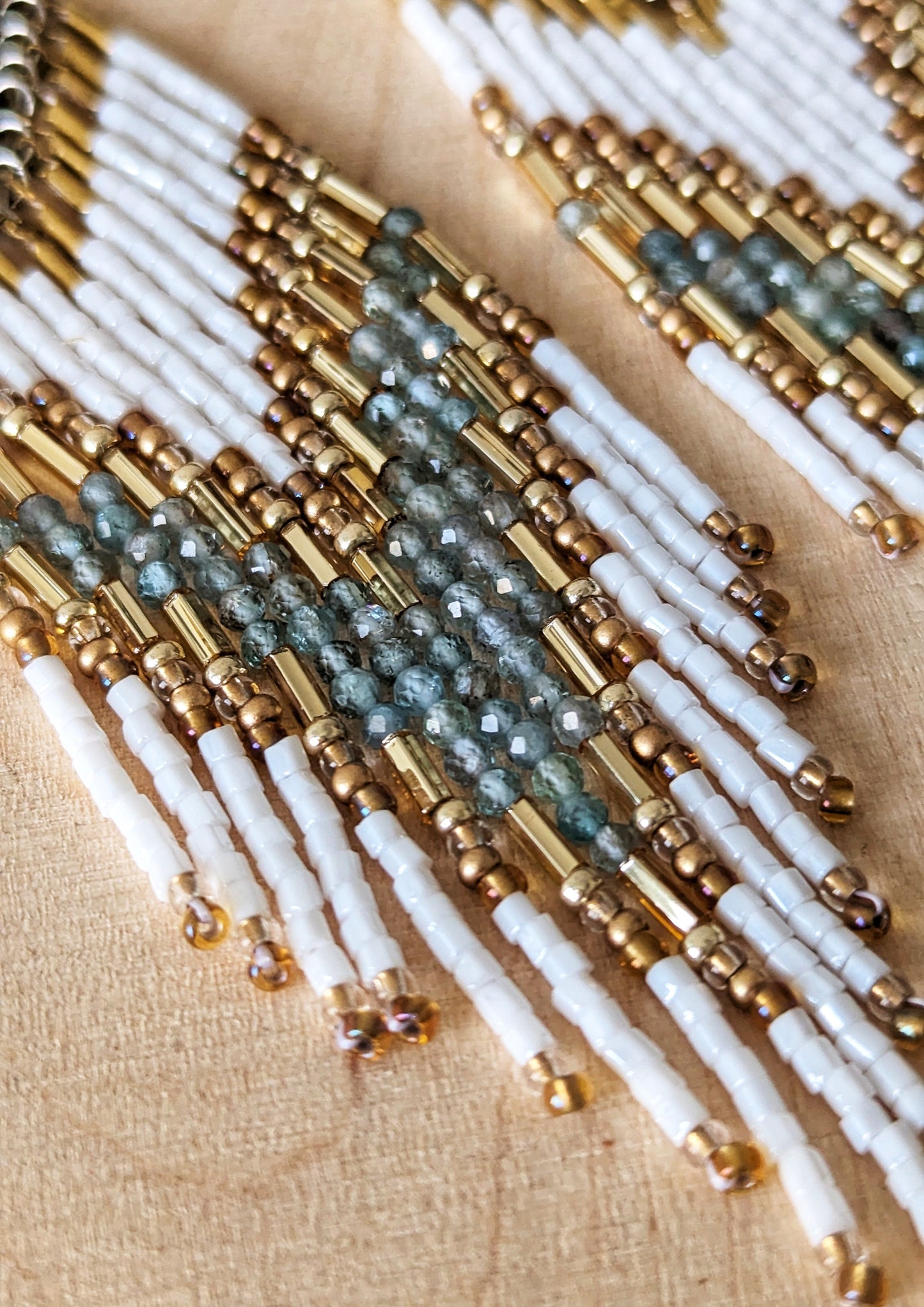 Elegant beaded earrings with white, gold, and soft aqua accents, displayed on a light wood background with delicate dried flowers nearby.- Moon & Milk