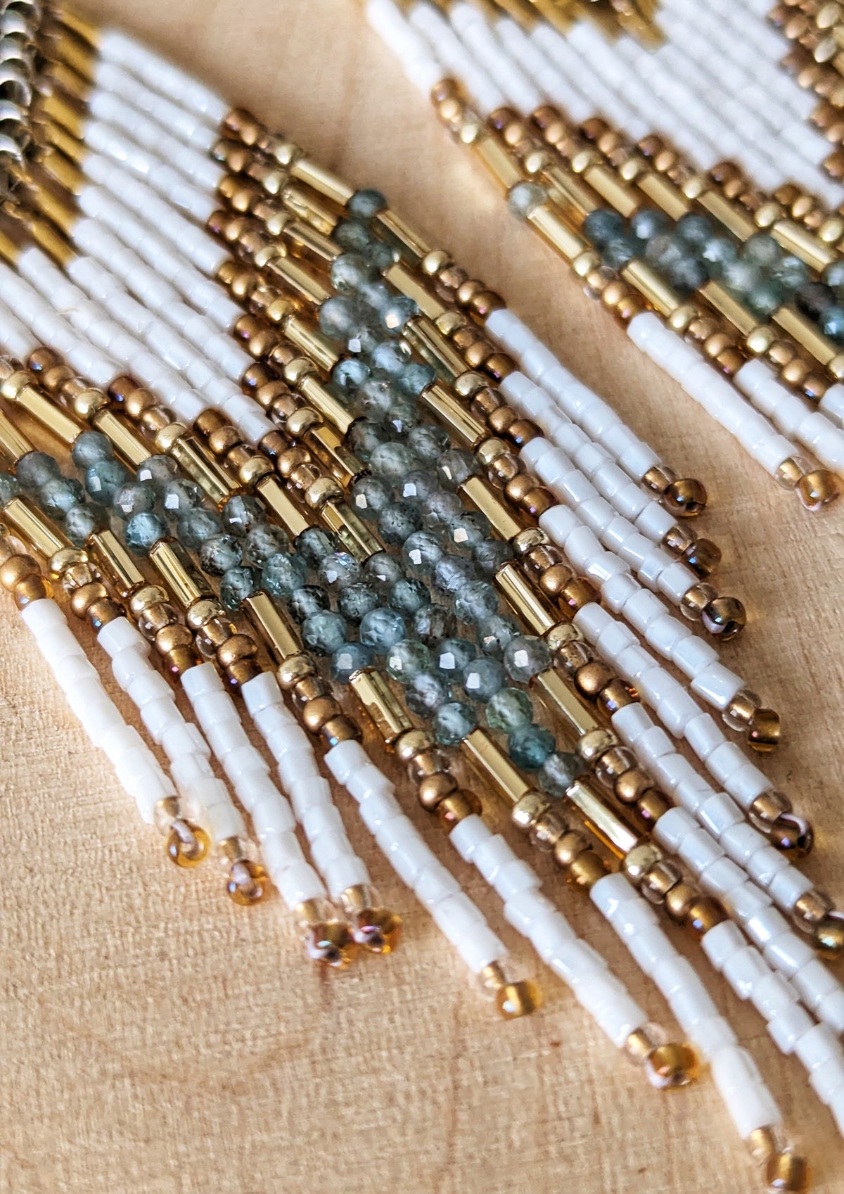 Elegant beaded earrings with white, gold, and soft aqua accents, displayed on a light wood background with delicate dried flowers nearby.- Moon & Milk