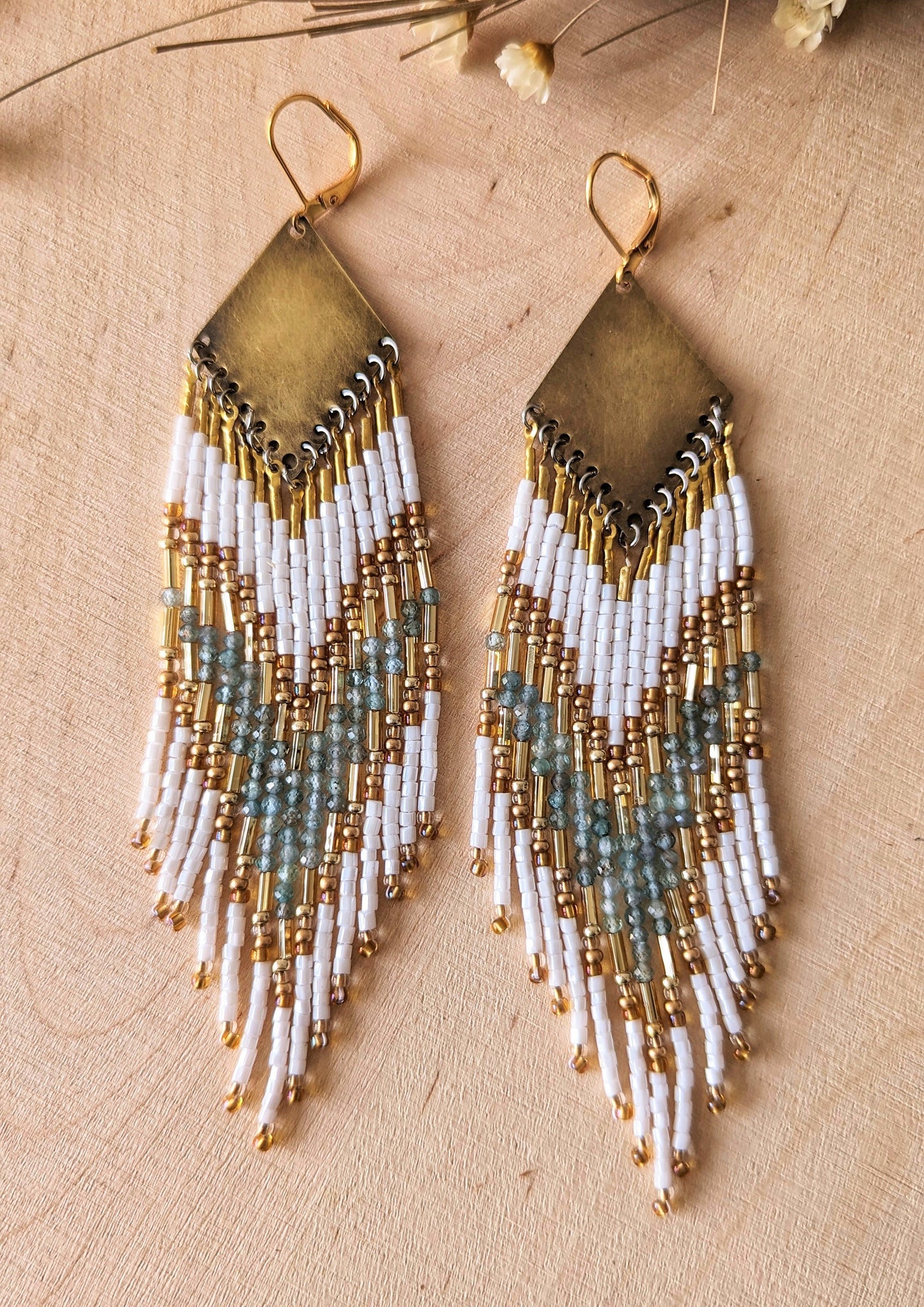 Elegant beaded earrings with white, gold, and soft aqua accents, displayed on a light wood background with delicate dried flowers nearby.- Moon & Milk