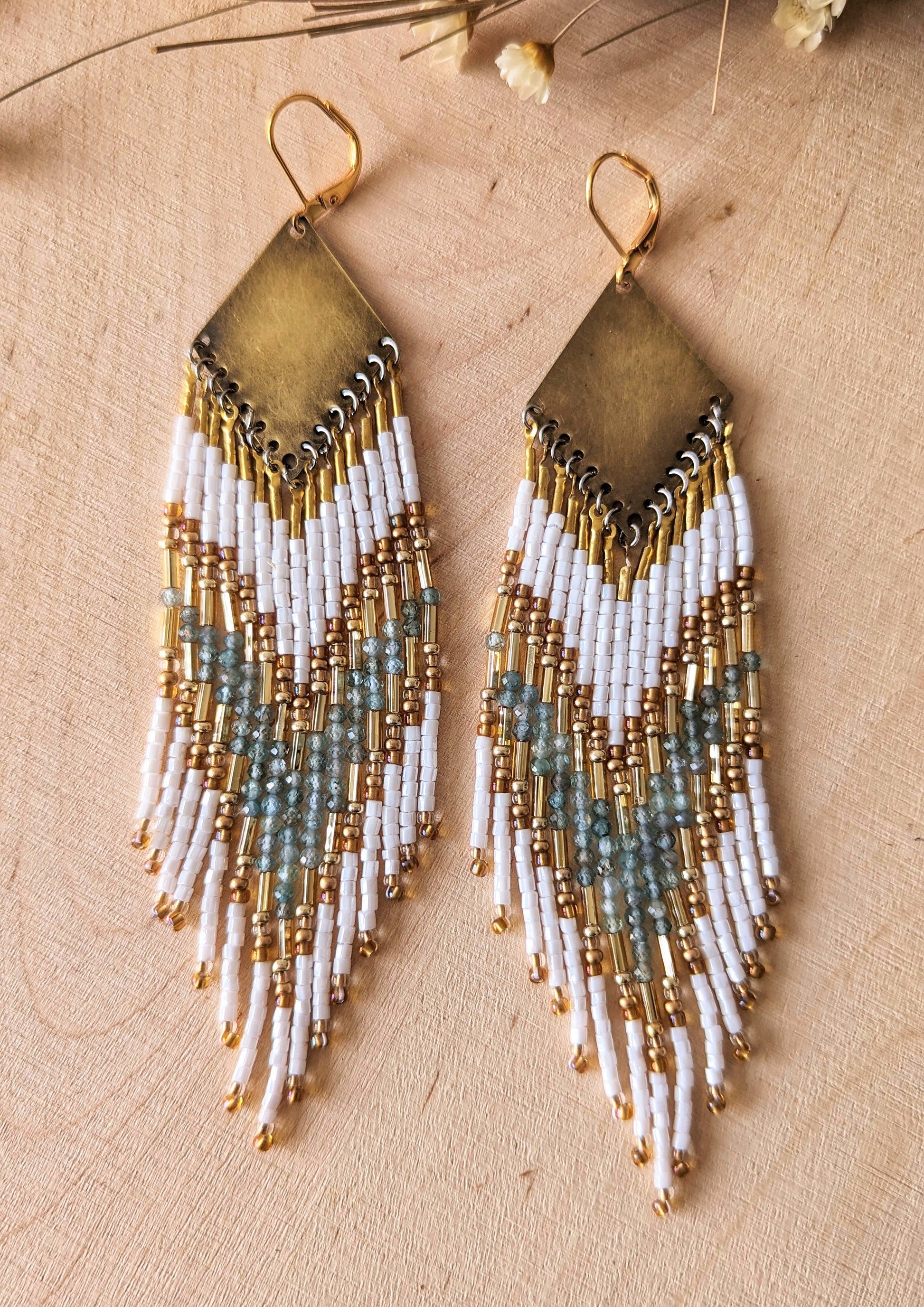 Elegant beaded earrings with white, gold, and soft aqua accents, displayed on a light wood background with delicate dried flowers nearby.- Moon & Milk