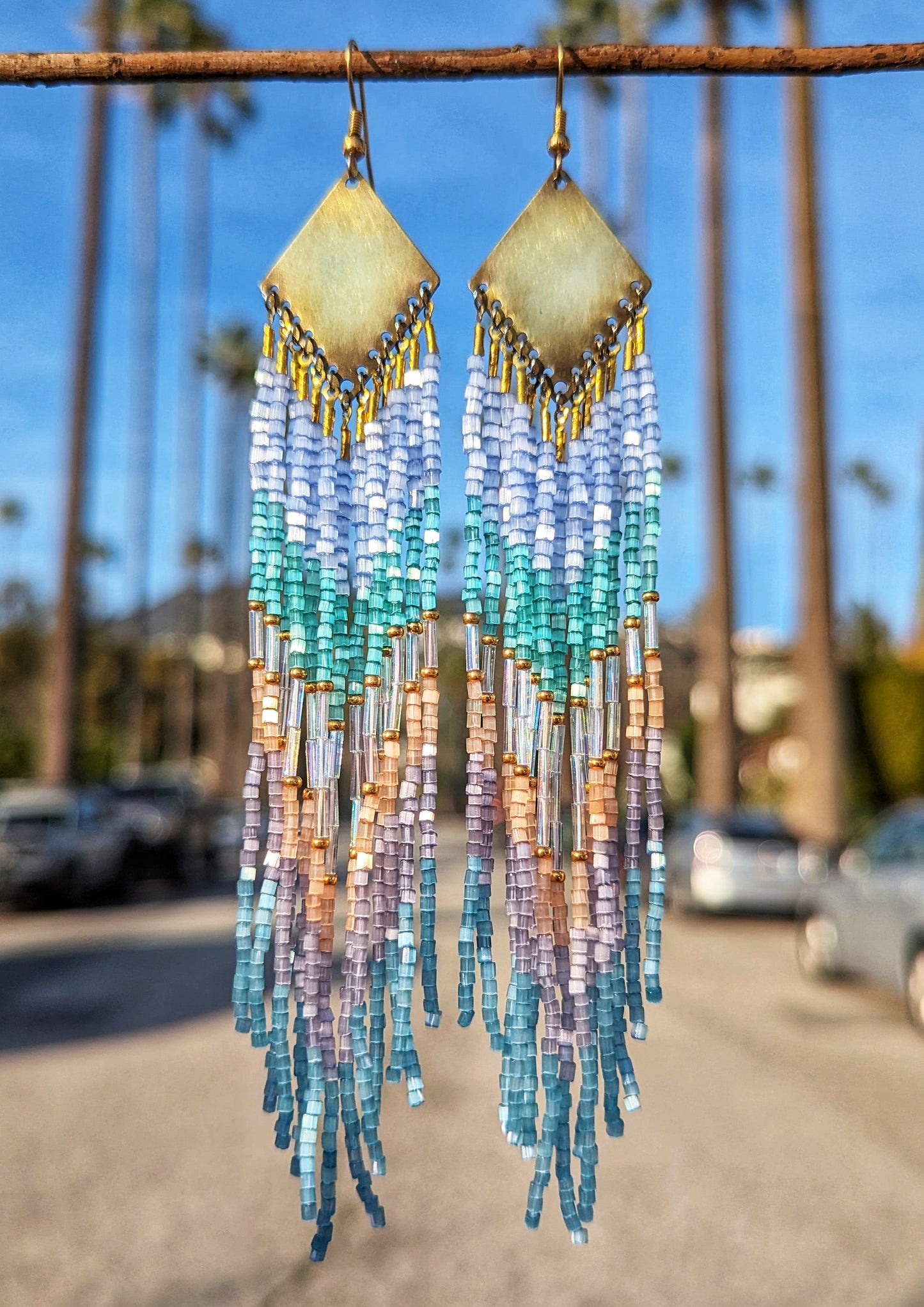 A pair of beaded earrings with brass accents and a variety of colors including blue and pink beads, displayed against an outdoor backdrop.
