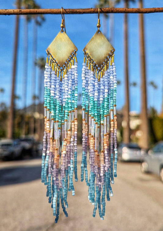 A pair of beaded earrings with brass accents and a variety of colors including blue and pink beads, displayed against an outdoor backdrop.