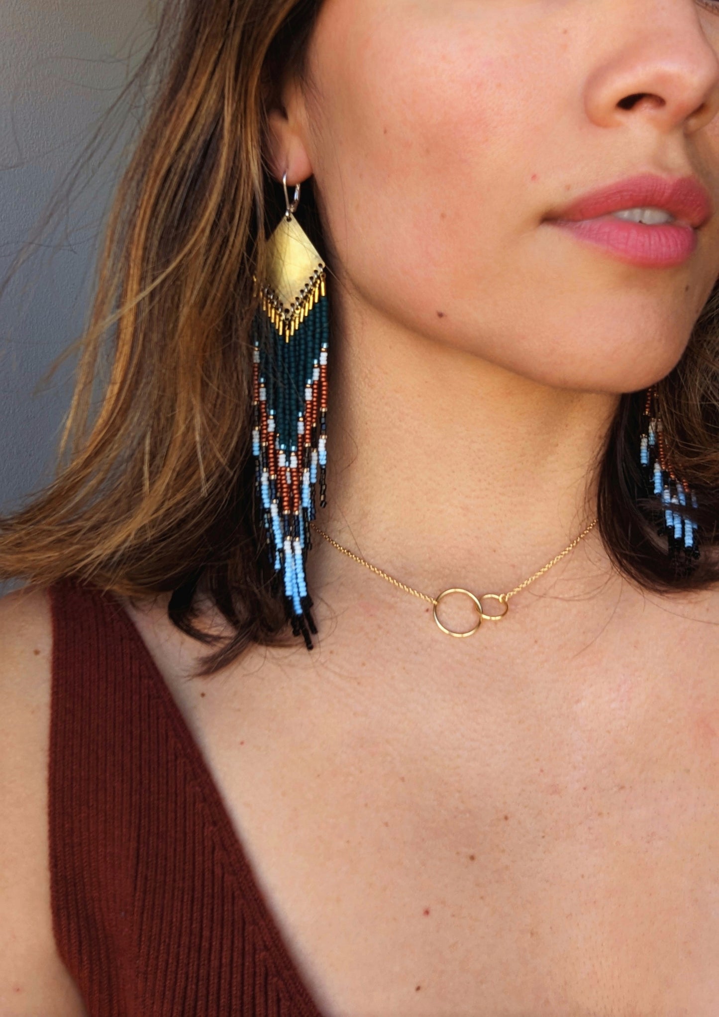 Beaded earrings with teal, rust, black, and ivory chevron pattern, styled with a brass top, worn by a model pairing it with a minimalist gold necklace.