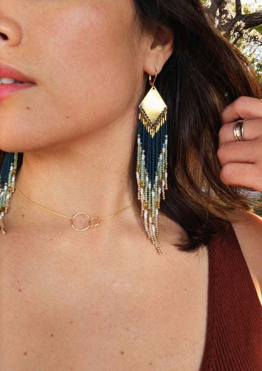 Close-up of a woman wearing Moon & Milk handcrafted beaded earrings with dark green, gold, and cream glass beads and brass accents, paired with a gold circle necklace.