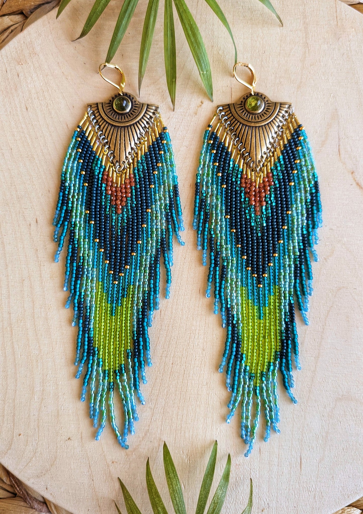 Pair of long beaded earrings featuring intricate patterns in deep blue, lime green, teal, and rust, with a brass sunburst accent at the top, and a dark green stone embedded, lying on a wooden surface.